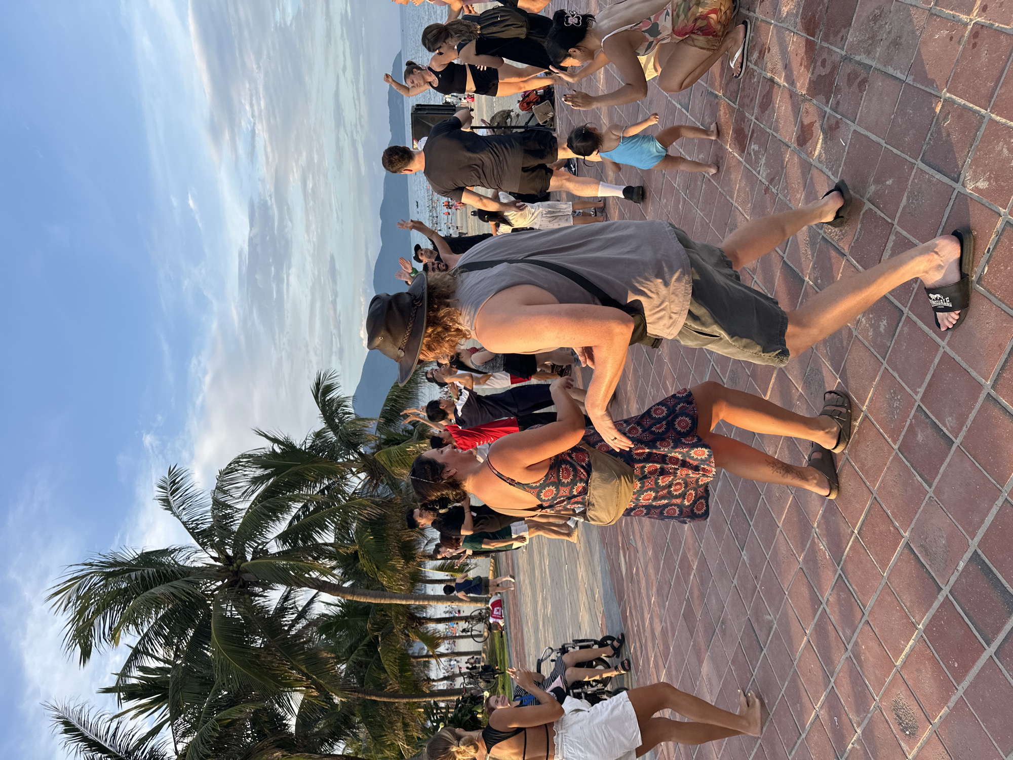This media file captures an outdoor group exercise session taking place on a paved promenade in Đà Nẵng, Vietnam, during the late afternoon or early evening. Numerous individuals of varying ages, including adults and children, are gathered, many actively engaged in what appears to be a yoga or stretching class. Participants are seen in various postures, some bending forward, others reaching upwards, following an unseen leader or common routine.
The scene is set against a tropical coastal backdrop. Lush palm trees line the promenade on the left, casting long shadows indicative of the time of day. In the background, beyond a sandy beach area, mountains rise under a clear blue sky dotted with wispy white clouds. The ground is paved with distinctive red-brown interlocking bricks or tiles.
In the foreground, a man in a grey t-shirt, shorts, and a hat is bending forward. Beside him, a woman in a floral dress is in a similar pose. A speaker system is visible in the midground, suggesting amplified instruction or music for the participants. Other details include people in casual attire, a child playing, and a person's black flip-flop with partially legible white text on its strap. The overall atmosphere appears focused yet relaxed, characteristic of a community fitness event.