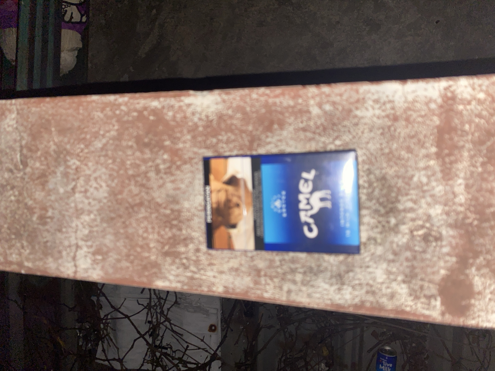 A pack of Camel cigarettes is lying on a reddish-brown textured surface that appears to be a railing or ledge. The cigarette pack is predominantly blue with white lettering and features the iconic Camel logo with a camel silhouette. In the background, there are dark shadows and hints of branches, suggesting an outdoor environment possibly with some foliage. A discarded can with faint blue and white lettering is visible on the lower left. The overall scene is dimly lit, contributing to a somewhat gritty atmosphere. The image does not contain any identifiable landmarks or contextual clues that would suggest a specific location within Tangerang, Indonesia.