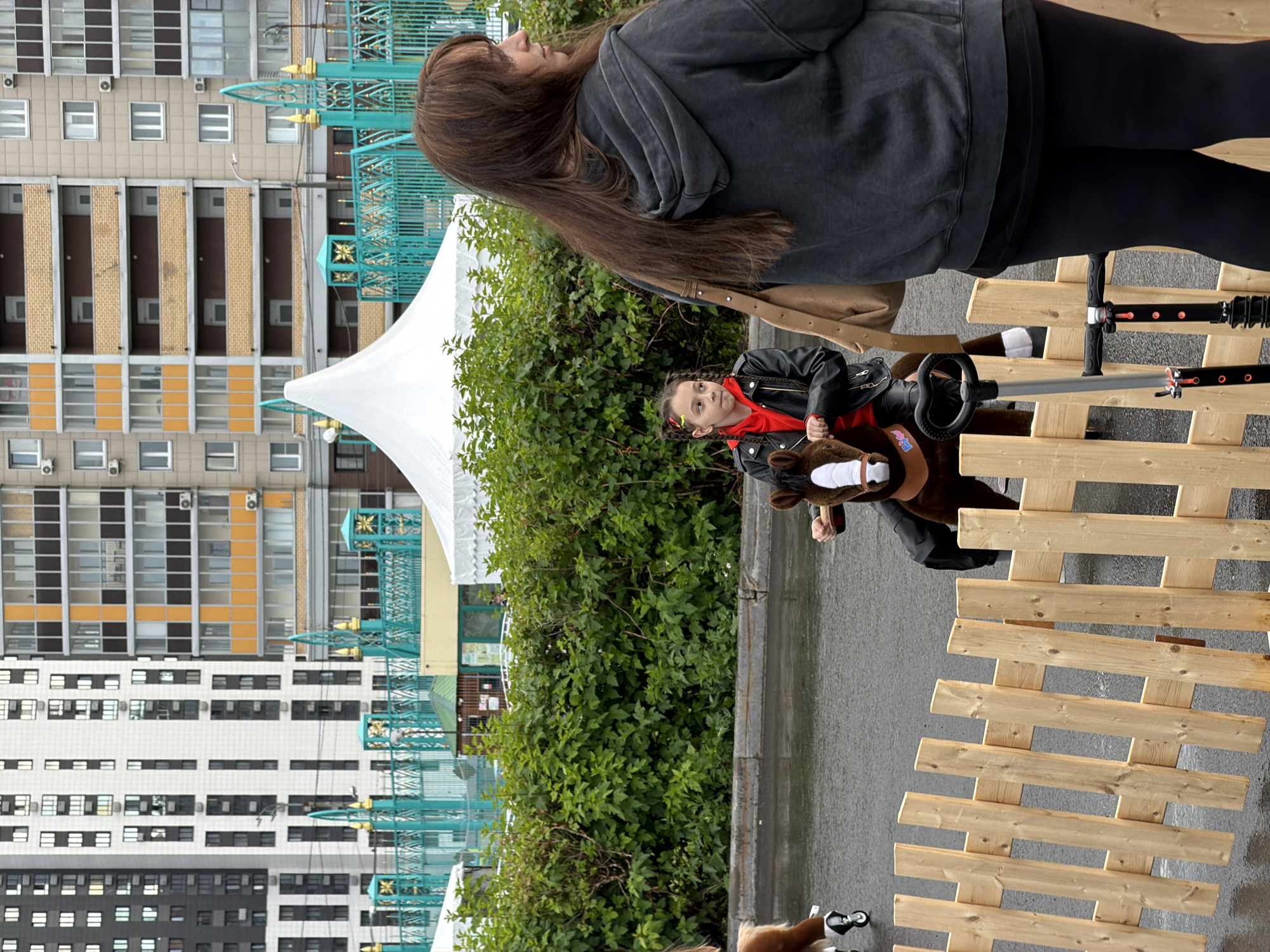 In a city environment, a young girl with braided hair, wearing a red hoodie and a black leather jacket, rides a toy horse with wheels. She is positioned behind a wooden fence, looking to her left. To her right, a woman with long brown hair, wearing a dark grey hooded sweatshirt and black leggings, stands facing away from the camera, also looking to her left. In the background, there are tall apartment buildings with numerous windows and balconies, as well as a white tent structure and green trees. The ground appears wet, suggesting recent rain. A scooter is visible near the fence.