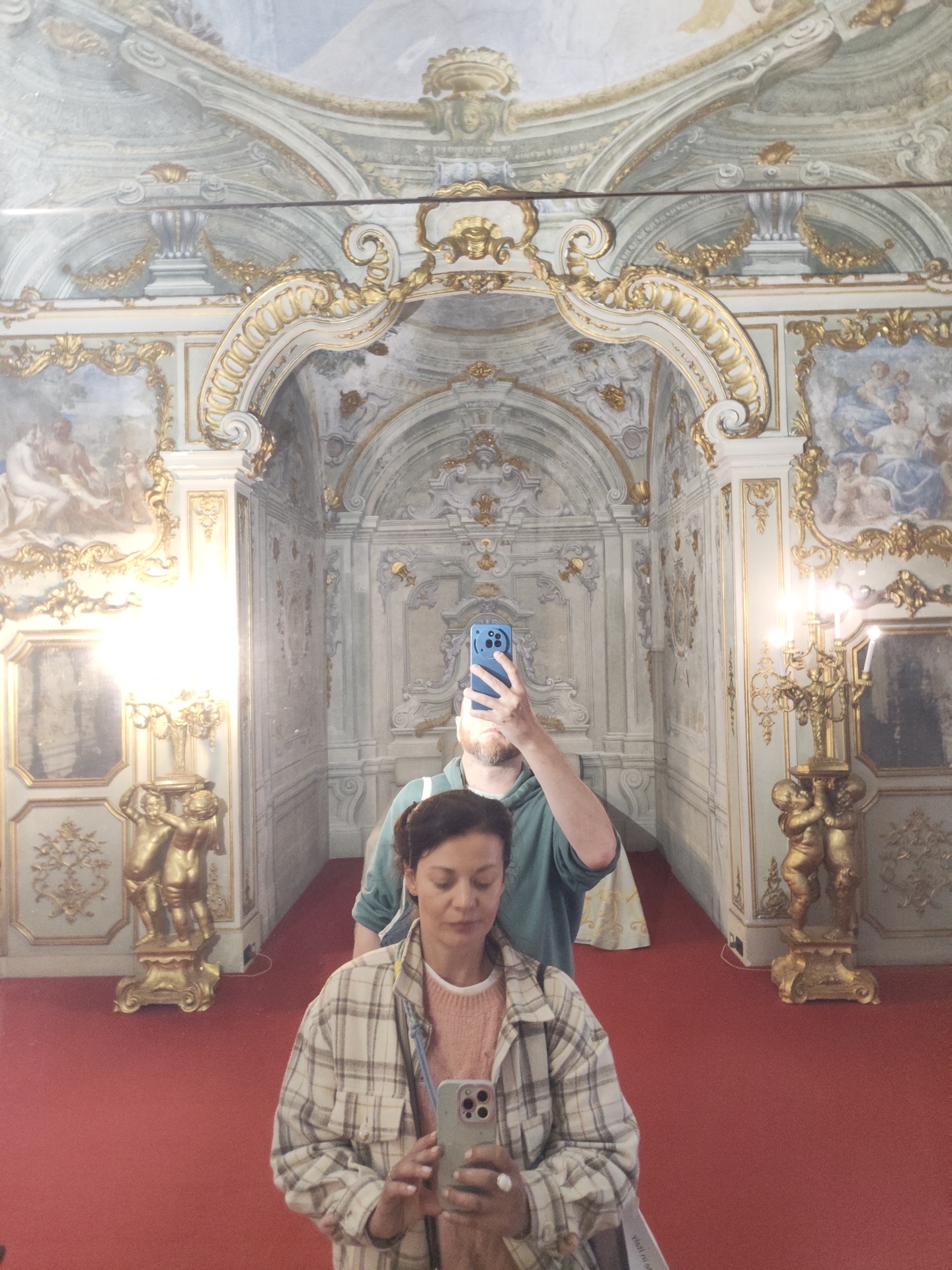 The image shows a mirrored reflection of a room in Palazzo Nicolosio Lomellino in Genoa, Italy. A woman in a tan and white plaid shirt and pink sweater is in the foreground, holding a smartphone and appearing to take a photo of herself in the mirror. Behind her, a man is holding a blue smartphone up to take a photo of the room.
The room's architectural style is ornate, featuring an archway decorated with white and gold details. The walls are adorned with paintings in elaborate frames. Golden statues stand on pedestals on either side of the room. The floor is covered with red carpeting. The ceiling of the room is painted with a fresco.