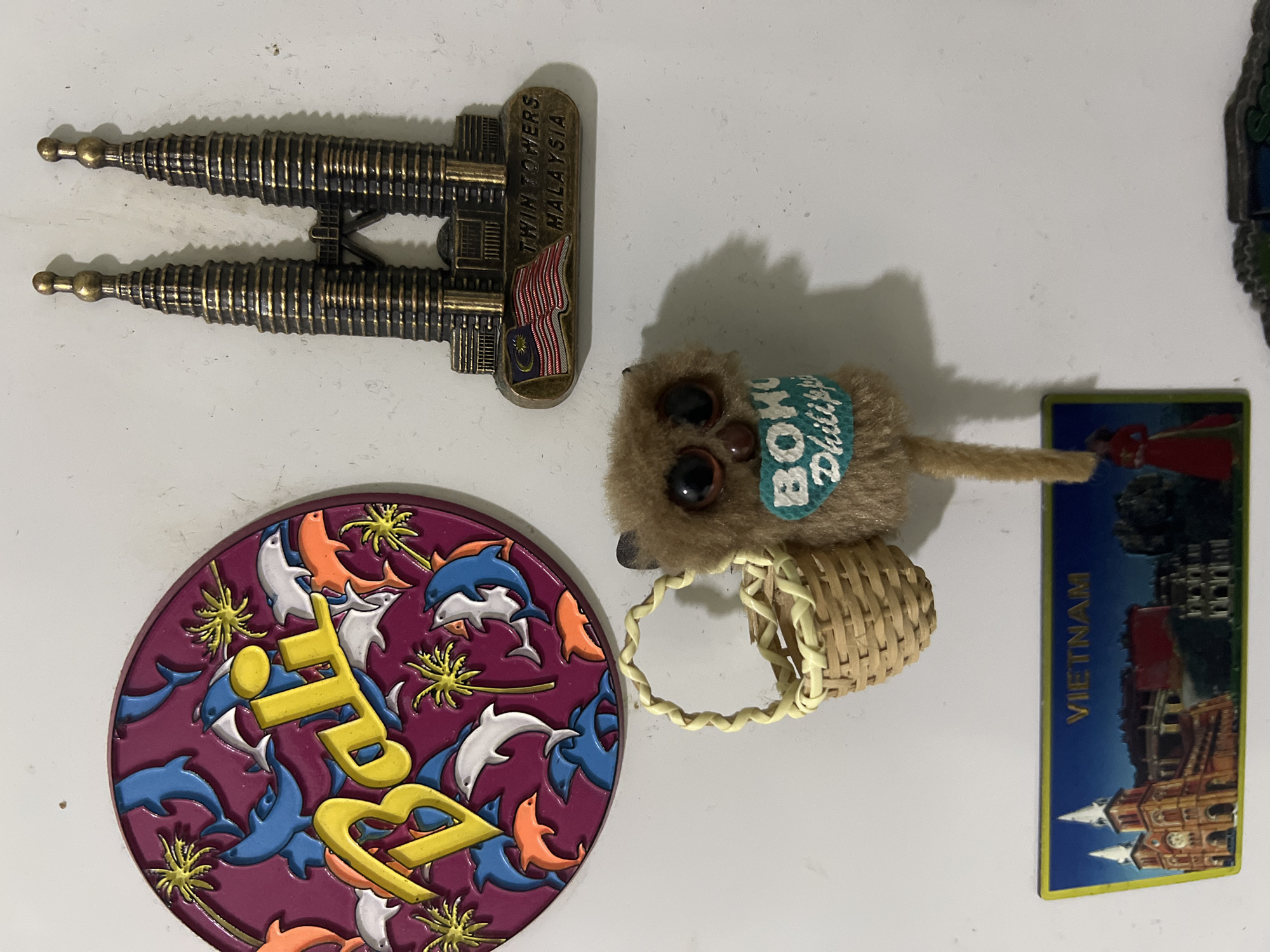 This media file contains a collection of souvenir magnets and a small plush toy, all placed on a white surface. The subjects are diverse and suggest travel or cultural interests.
In the top left, there is a magnet shaped like the Petronas Twin Towers in Malaysia, with the text "TWIN TOWERS, MALAYSIA" and a depiction of the Malaysian flag. To its right is a small, fuzzy plush toy with large, dark eyes. It wears a teal band around its neck with the words "BOKO, PHILIPPINES" visible. The toy also holds a miniature woven basket.
Below the Twin Towers magnet is a round, purple magnet featuring a design of leaping dolphins and palm trees, with the word "BALI" prominently displayed in yellow letters.
To the right of the plush toy, there is a rectangular magnet with a blue background and an image of a grand building, likely a historical or religious structure, with the word "VIETNAM" written vertically in yellow text.
The overall scene is an arrangement of these items on a plain white background, suggesting they might be displayed together, perhaps on a refrigerator or a similar magnetic surface. There is no indication of people, specific time of day, weather, or emotions, beyond the inherent appeal of travel souvenirs. The location context of Malé, Maldives, does not appear to be directly represented by any of the visible objects or text within the image itself.