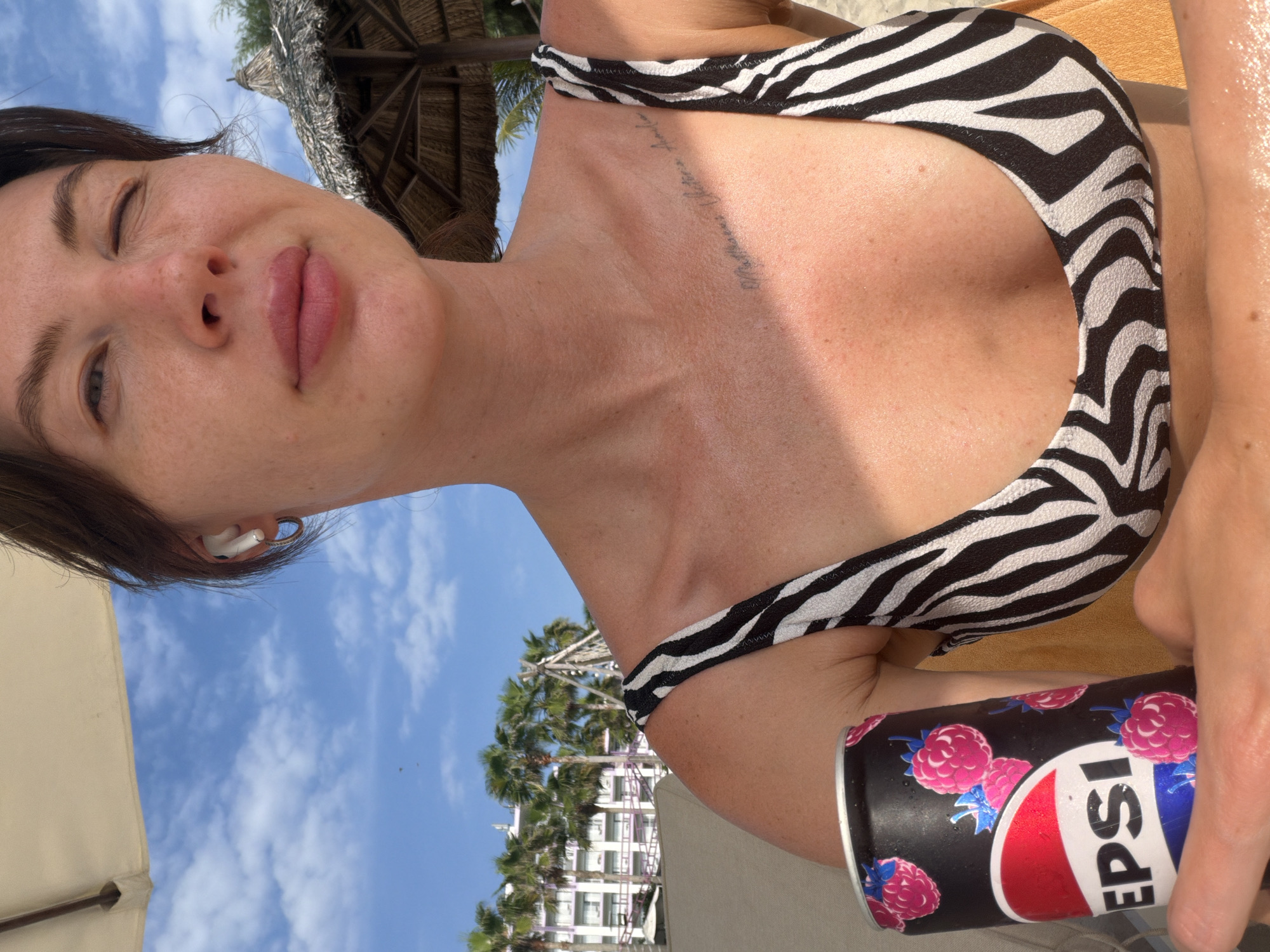 A woman with dark hair is holding a can of Pepsi. She is wearing a black and white zebra-print bikini top. The background shows palm trees and a building, suggesting a tropical resort. The sky is blue with scattered clouds. The woman is partially in the frame, with her face turned towards the camera, and she appears to be winking. A tattoo is visible on her collarbone. The Pepsi can has a design of raspberries. The scene is set outdoors, likely on a beach or near a pool. The specific location is Dong Ninh Hoa Ward, Vietnam.