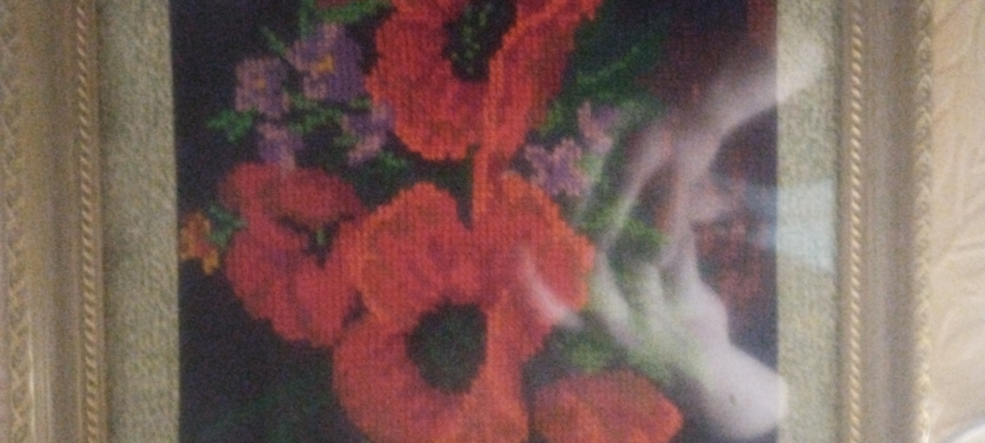 This is a close-up, high-angle, blurry photograph of a framed needlepoint artwork featuring a bouquet of poppies and other flowers. The poppies are a vibrant red, with dark centers, and are the most prominent elements in the composition. There are also smaller, purple flowers interspersed with the poppies. The background is a dark, muted color, which makes the red of the poppies stand out. The artwork is framed by a decorative gold and beige frame. The image quality is poor, with a significant amount of noise and blur, making it difficult to discern fine details of the needlepoint. There are no indications of a specific location within a city.