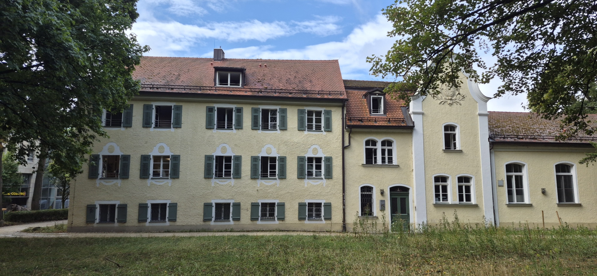 The image shows a pale yellow, two-story building with a red-tiled roof in Regensburg, Germany. The building has multiple windows, some with green shutters, and architectural embellishments above some of the upper-story windows. A portion of the building to the left, which is taller and also has a red-tiled roof, is connected to the main structure. Trees with green foliage are visible in the foreground and framing the right side of the building. The ground in front of the building is covered in dry grass. The sky above is blue with scattered clouds.