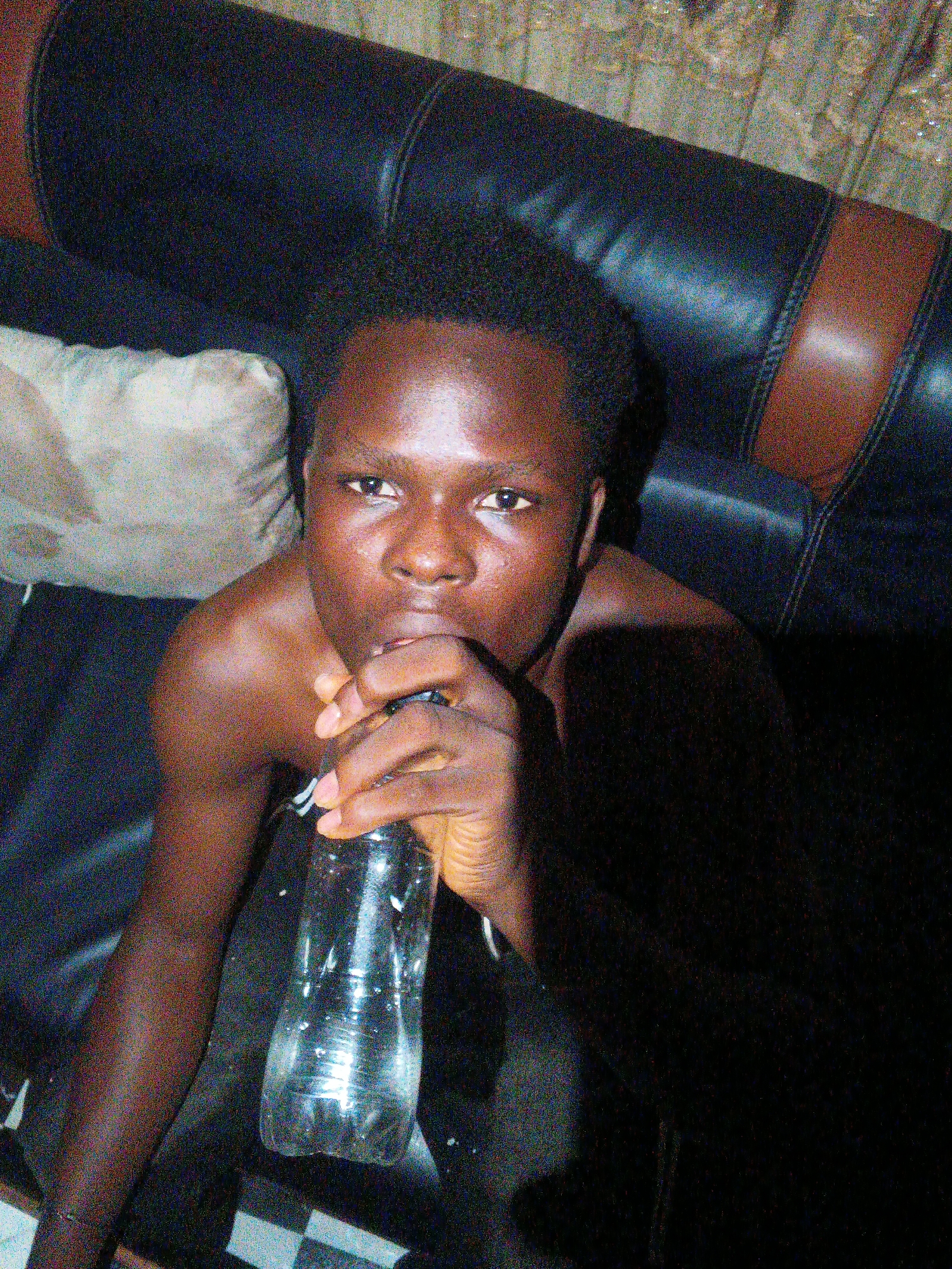 A young, shirtless Black man is drinking from a clear plastic water bottle. He is sitting on a dark leather couch with a tan leather accent. To his left is a beige pillow. The photo is taken from an elevated angle. The lighting is poor and the image grainy. The location, Onitsha, Nigeria, is not visually verifiable from the image.