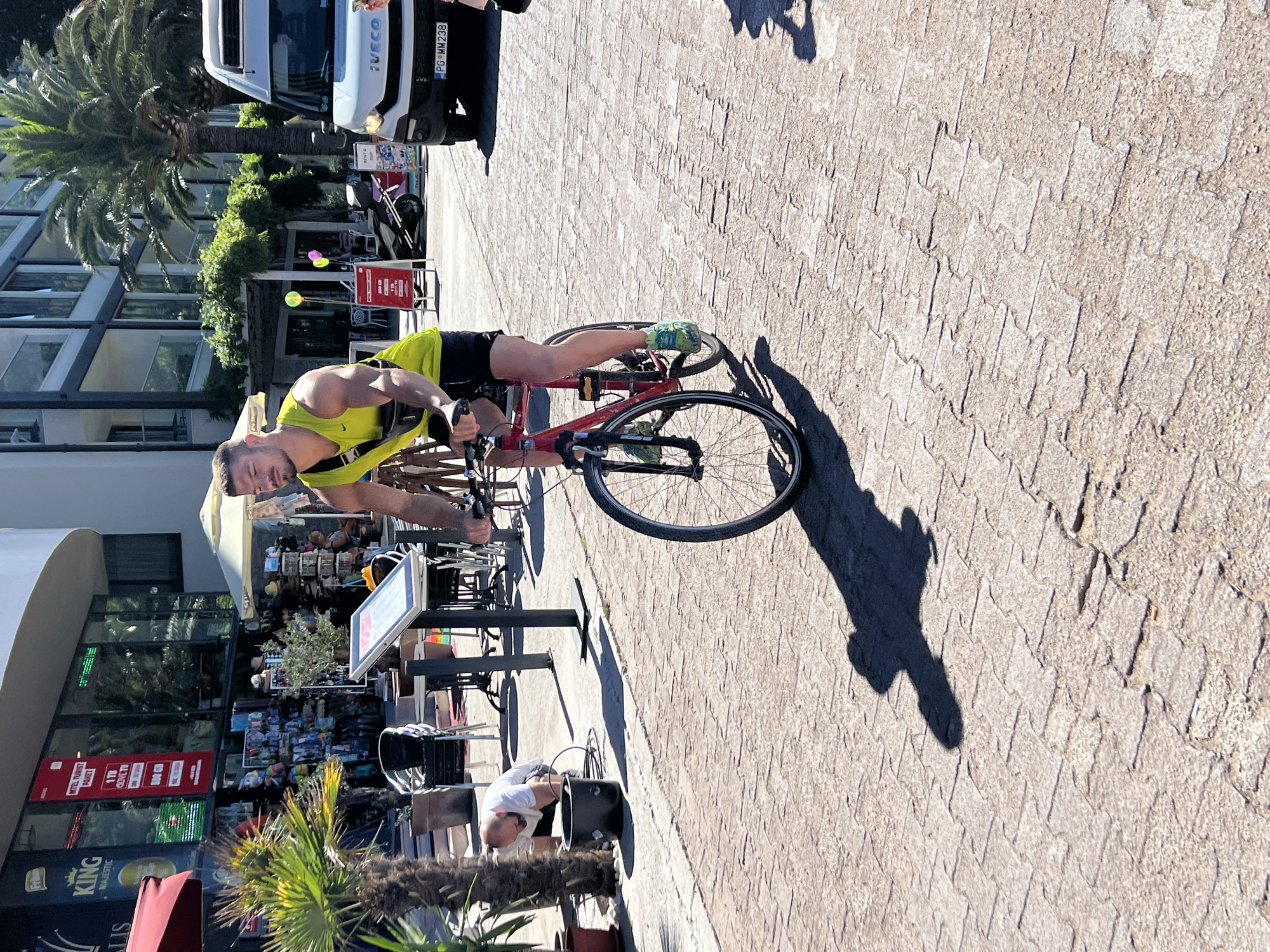A man is riding a red bicycle on a paved walkway in the city of Rafailovići, Montenegro. He is wearing a bright yellow sleeveless top and dark shorts, with a black bag slung across his chest. The bicycle has a black frame and black wheels. To the left of the cyclist, a man in a white shirt and dark shorts is bent over a black bucket, with a hose running into it. Behind them is a building with multiple floors, palm trees, and shops displaying various goods. A white van is parked on the right side of the image. The walkway is made of interlocking cobblestones, and the scene is illuminated by bright sunlight, casting distinct shadows.