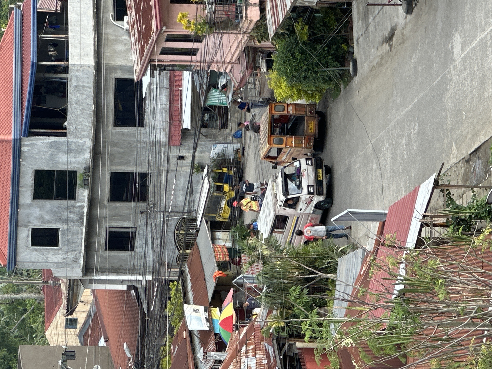 The image shows a street scene in Davao City, Philippines. The view is from an elevated position, looking down on a narrow street with buildings on either side. The buildings are a mix of concrete structures and dwellings with corrugated metal roofs, typical of a densely populated urban area. Many electrical wires are visible. Several jeepneys, public transportation vehicles, are present on the street. People are walking and standing near the jeepneys. The general environment is urban and densely populated.