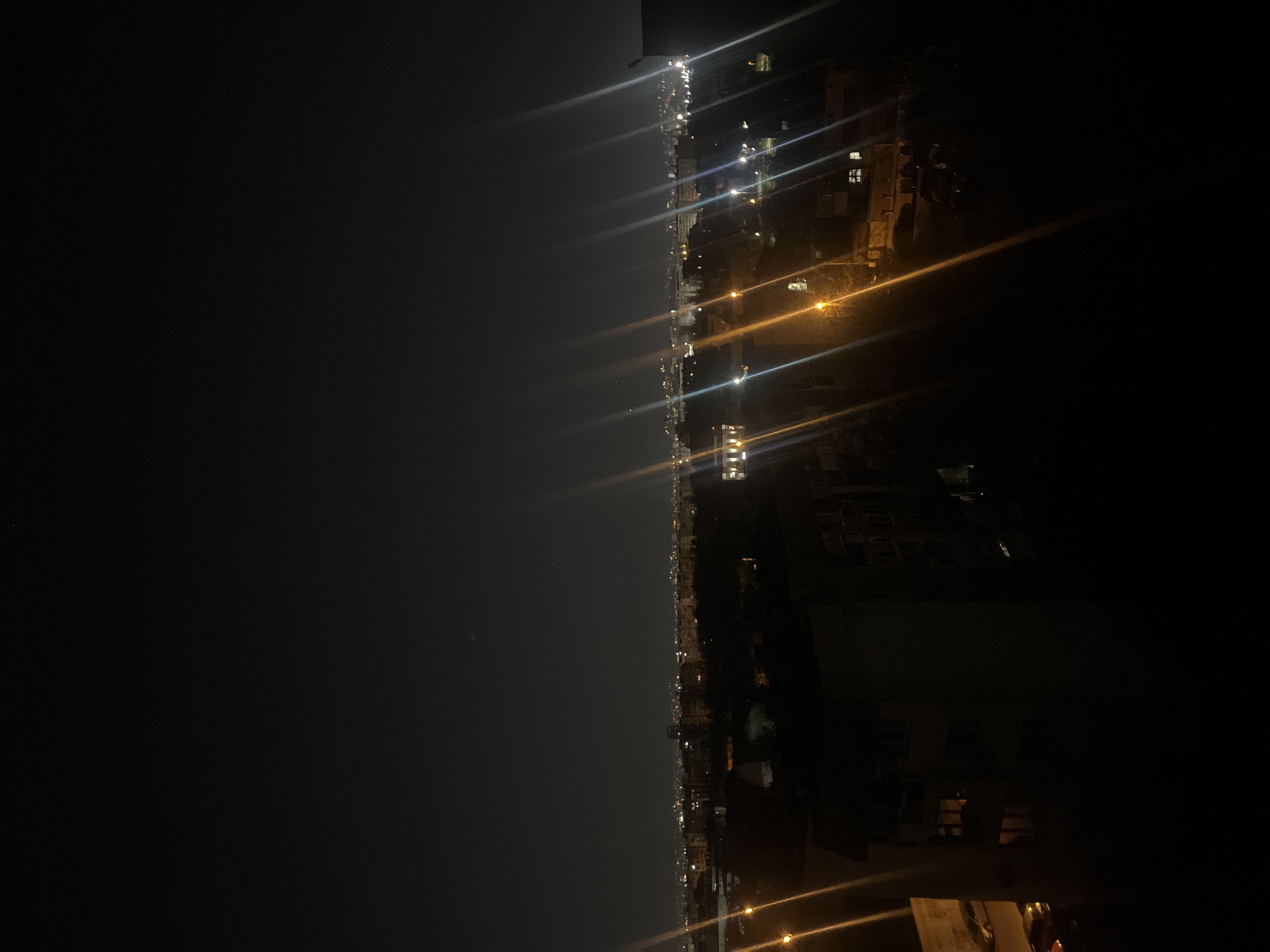 This is a nocturnal photograph capturing an expansive view of a city, likely Sofia, Bulgaria, from an elevated vantage point. The image depicts a vast expanse of dark night sky on the left, transitioning to an illuminated urban landscape on the right side of the frame.
The cityscape is characterized by numerous artificial lights, predominantly from buildings and streetlights, which create a glittering tapestry across the horizon. Many of these light sources produce pronounced lens flares or streaks that emanate outwards, adding a dynamic visual effect to the scene. The lights exhibit a range of colors, including bright whites, cool blues, and warm yellows and oranges. Dark, silhouetted forms of buildings and other urban structures are faintly visible amidst and below the clusters of lights, hinting at the dense development of the city. The horizon line is discernible, indicating the extensive spread of the city's illumination.
The time of day is clearly night, evidenced by the darkness of the sky and the reliance on artificial illumination. The sky appears clear, without significant cloud cover. There are no people visible, nor are there any specific activities or events taking place that can be identified. No visible text is present in the image. The photograph offers a sense of the scale and nocturnal ambiance of an urban environment.