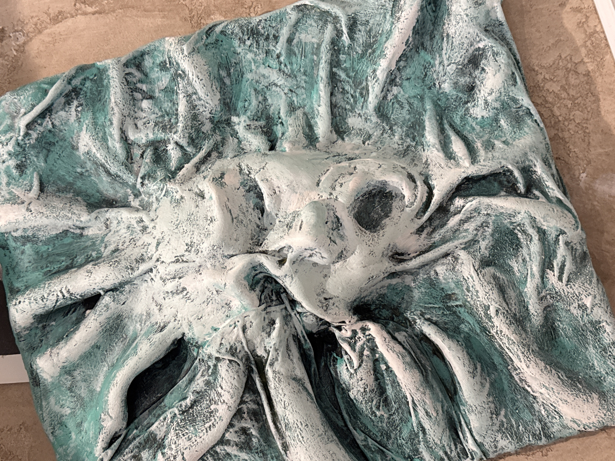 The image shows a close-up view of a textured, relief sculpture of a face with swirling, wave-like forms radiating outwards. The sculpture is painted in a distressed style with white and teal colors, giving it an aged or weathered appearance. The face appears to be contorted in an expression of anguish or surprise, with an open mouth and closed eyes. The background is a textured, beige surface, possibly a wall or floor. There is no discernible location in Sofia, Bulgaria, visible in this image.