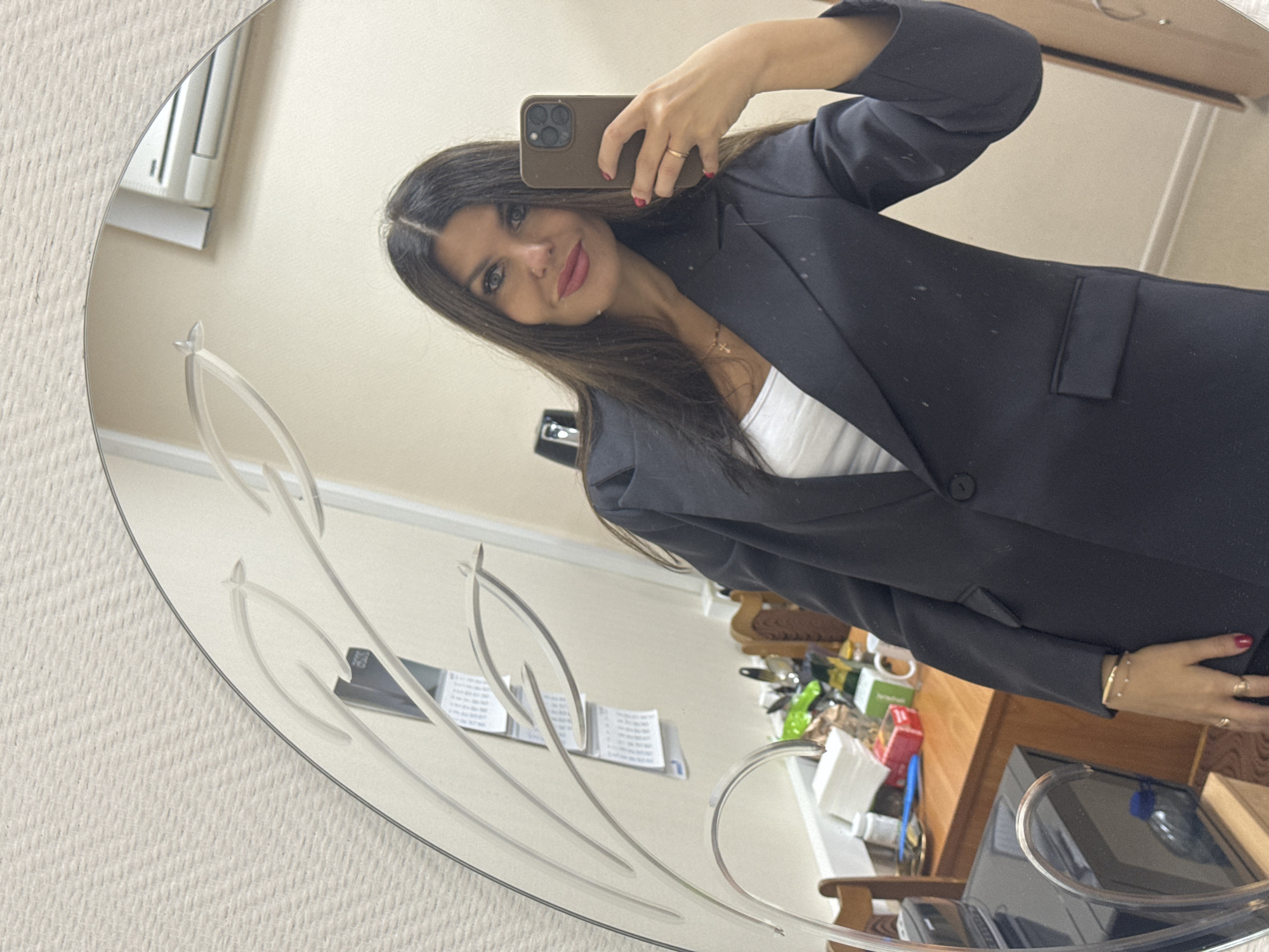 A woman is taking a selfie in a mirror. She is wearing a black blazer over a white top and has long dark hair. She is wearing jewelry on her neck, wrist, and fingers. Her nails are painted red. The mirror has an etched design of leaves. In the reflection, a desk with various items is visible, including a calendar, mugs, and containers. Behind her, a wall with an air conditioning unit is visible. The environment appears to be an indoor office or home setting. There is no visible indication of the city of Chaikovsky, Russia in the image.