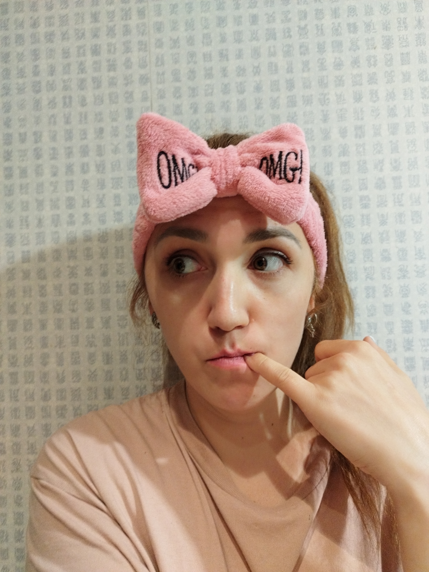 The image shows a young woman with brown hair, wearing a pink headband with the embroidered text "OMG!". She is looking to her left with her finger touching her lower lip, suggesting a contemplative or surprised expression. The background is a wall with a repeating pattern of grey geometric shapes. The woman is wearing a light pink t-shirt. The scene appears to be indoors, likely in a residential setting. There is no identifiable information in the image that would indicate a specific location within Saint Petersburg, Russia.