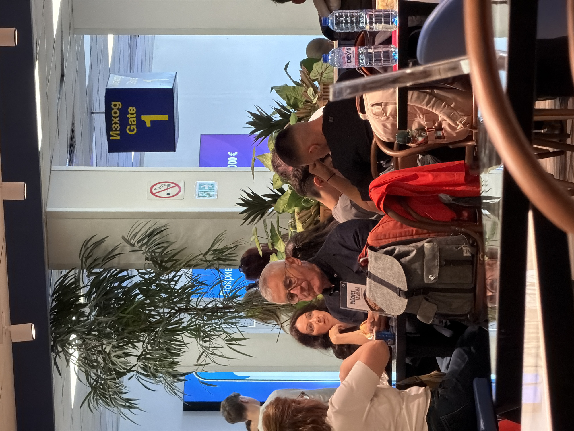The image shows a group of people sitting at tables in what appears to be a restaurant or cafe located in the Sofia Airport (SOF) in Bulgaria. A blue sign indicates "Gate 1." The group is diverse, with people of varying ages, hair colors, and clothing. Some are sitting, and others might be engaged in conversations. On the tables, there are items like water bottles, bags, and what may be food menus. Decorations such as plants add to the ambiance of the indoor setting. The environment seems like a typical waiting area in an airport, likely near a boarding gate.