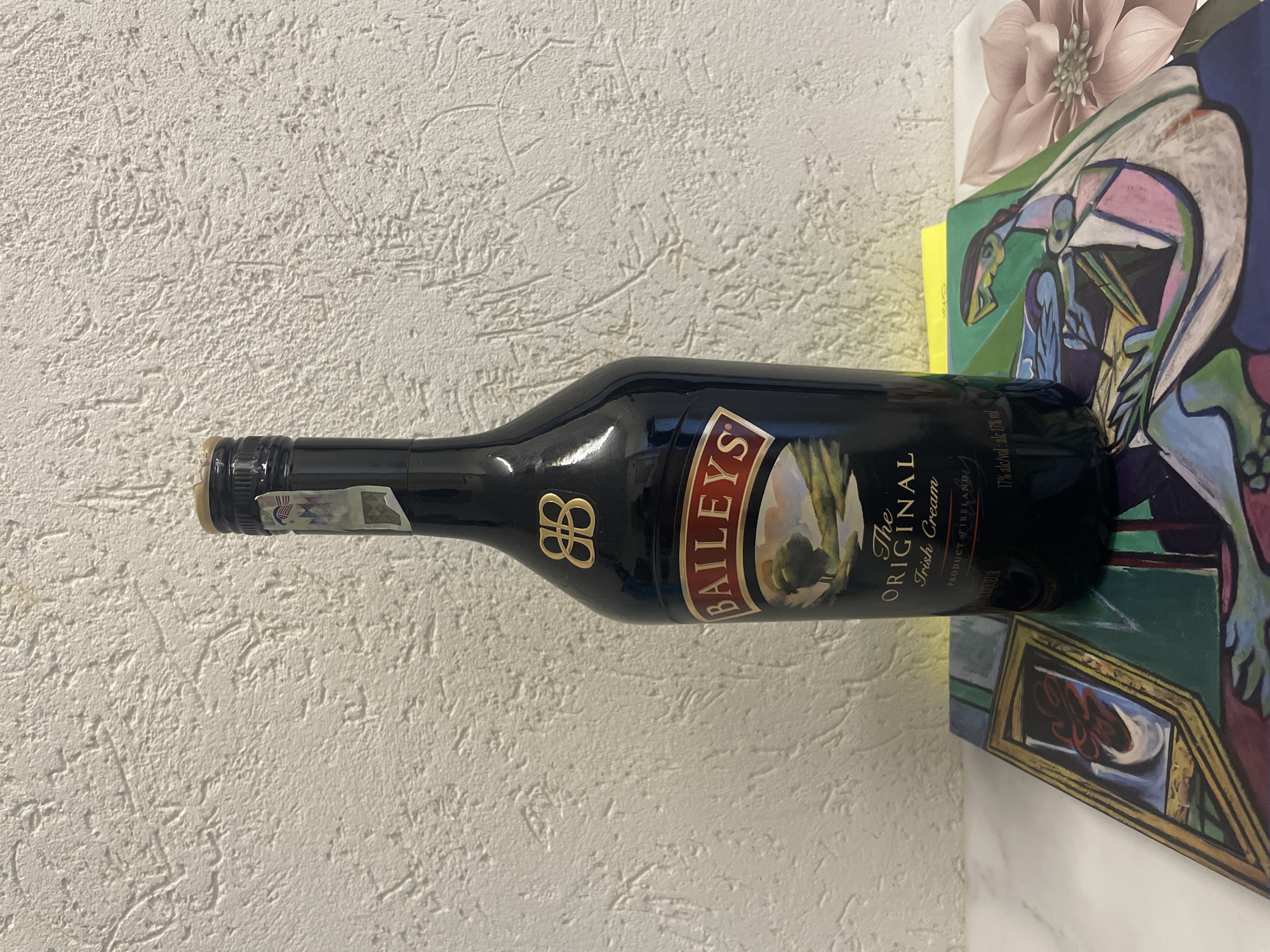 A horizontal eye-level indoor shot depicts a bottle of Baileys Irish Cream liqueur. The bottle is positioned against a textured, light-colored wall, and part of a painting is visible to the right of the bottle. The liqueur bottle is dark brown, with the brand name "BAILEYS" prominently displayed on the label in an arched, tan-colored font. Below the brand name, the label states "The ORIGINAL Irish Cream" in smaller lettering, followed by "PRODUCT of IRELAND" and "LIQUEUR". The alcohol content "17% alc/vol: alc 17% vol" is printed near the bottom right of the bottle. The bottle is sealed with a dark cap and has a small sticker on the neck. The painting behind the bottle features abstract figures with colors like blue, green, pink, and yellow. The scene is a generic indoor setting and cannot be specifically placed in Sofia, Bulgaria without additional context.