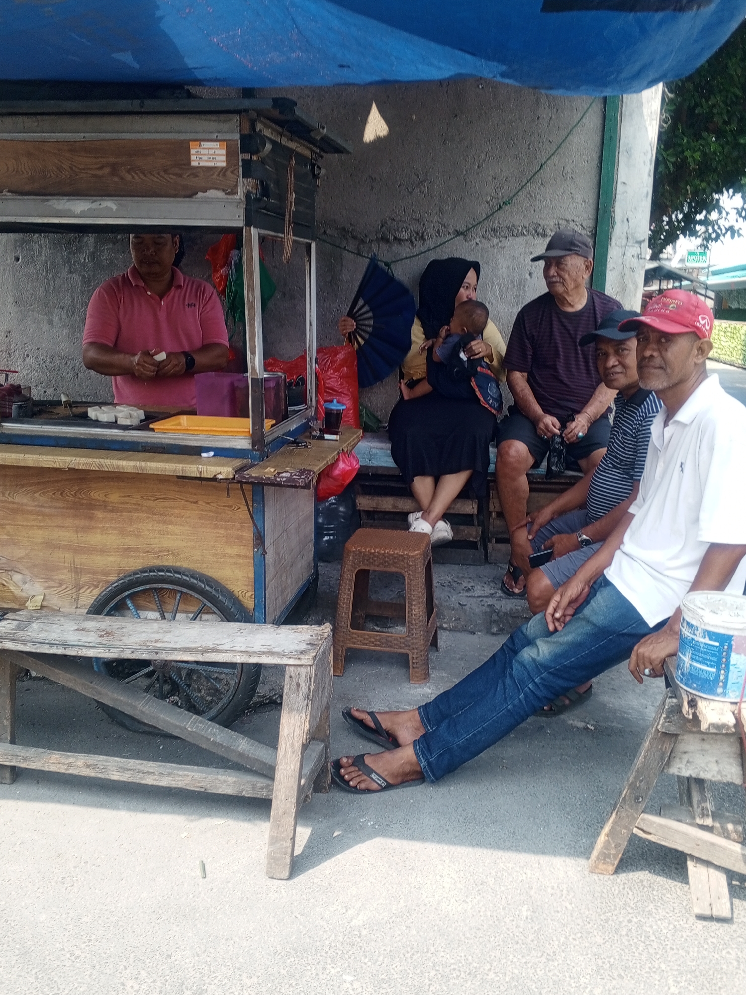 The image depicts a street food stall in Depok, Indonesia, under a blue tarp. A vendor in a pink polo shirt is preparing food behind the stall, which has a wooden front and a metal roller door. Several people are gathered around the stall, some sitting on wooden benches and stools. A woman wearing a hijab is holding a baby, and an elderly man wearing a cap is seated next to her. Two other men are also present, one wearing a striped shirt and a cap, and another in a red cap and white polo shirt. The environment appears to be a street or sidewalk, with a concrete surface and a wall in the background. The scene suggests a casual gathering of people interacting with the street vendor.