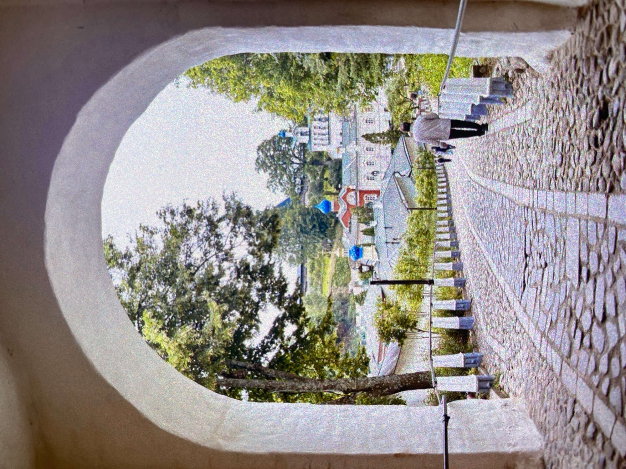 The image depicts a view through a stone archway, likely part of a monastery or similar structure. Beyond the archway, a paved pathway lined with low stone walls and vegetation leads towards a cluster of buildings with distinctively colored domes, a common characteristic of Russian Orthodox churches. A person is seen walking along the path. The location is likely within the city of Saint Petersburg, Russia, given the architectural style of the buildings and the typical urban landscape.