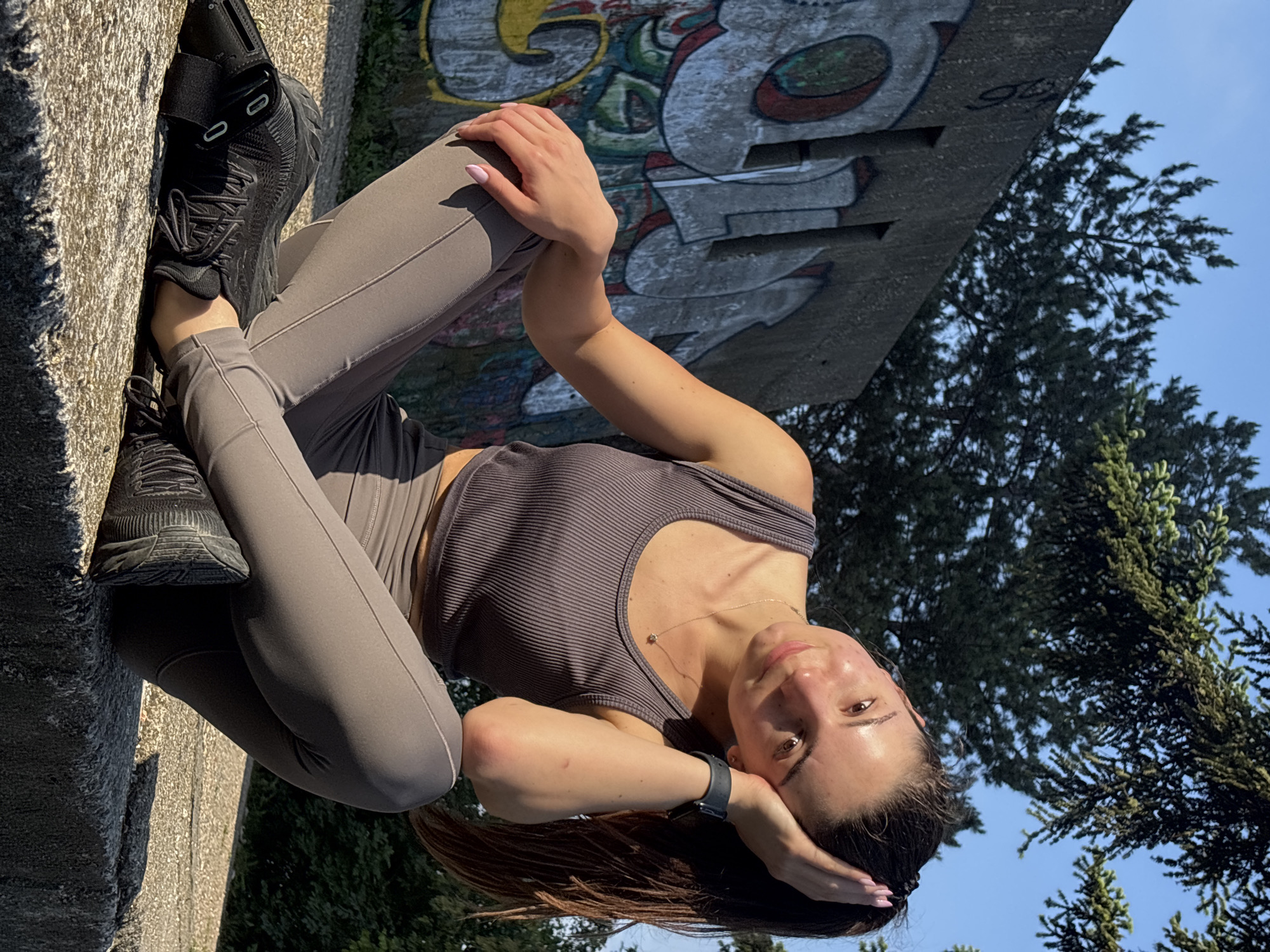 A young woman is captured in an outdoor setting, likely Varna, Bulgaria, in a photo rotated 90 degrees counter-clockwise. She is positioned against a rough, textured concrete structure, appearing to lean or lie back. She wears a casual, athletic outfit consisting of a dark grey ribbed tank top, grey form-fitting leggings, and black sneakers with visible straps and laces. A black smartwatch is on her left wrist, and a delicate gold necklace adorns her neck. Her fingernails are painted a light pink.
The woman gazes towards the camera with a calm, gentle expression, a slight smile on her lips. Her dark hair is loose, and one hand is resting near her head. The scene is brightly lit by natural sunlight, indicating a clear, sunny day.
Behind the concrete structure, a vibrant graffiti mural featuring abstract shapes and stylized letters in various colors including grey, red, yellow, and green can be seen, though the specific text is largely illegible. Further in the background, lush green trees are visible against a clear blue sky, suggesting an outdoor environment such as a park or urban green space. The composition creates a dynamic interplay between the subject, the urban concrete, and the natural elements.