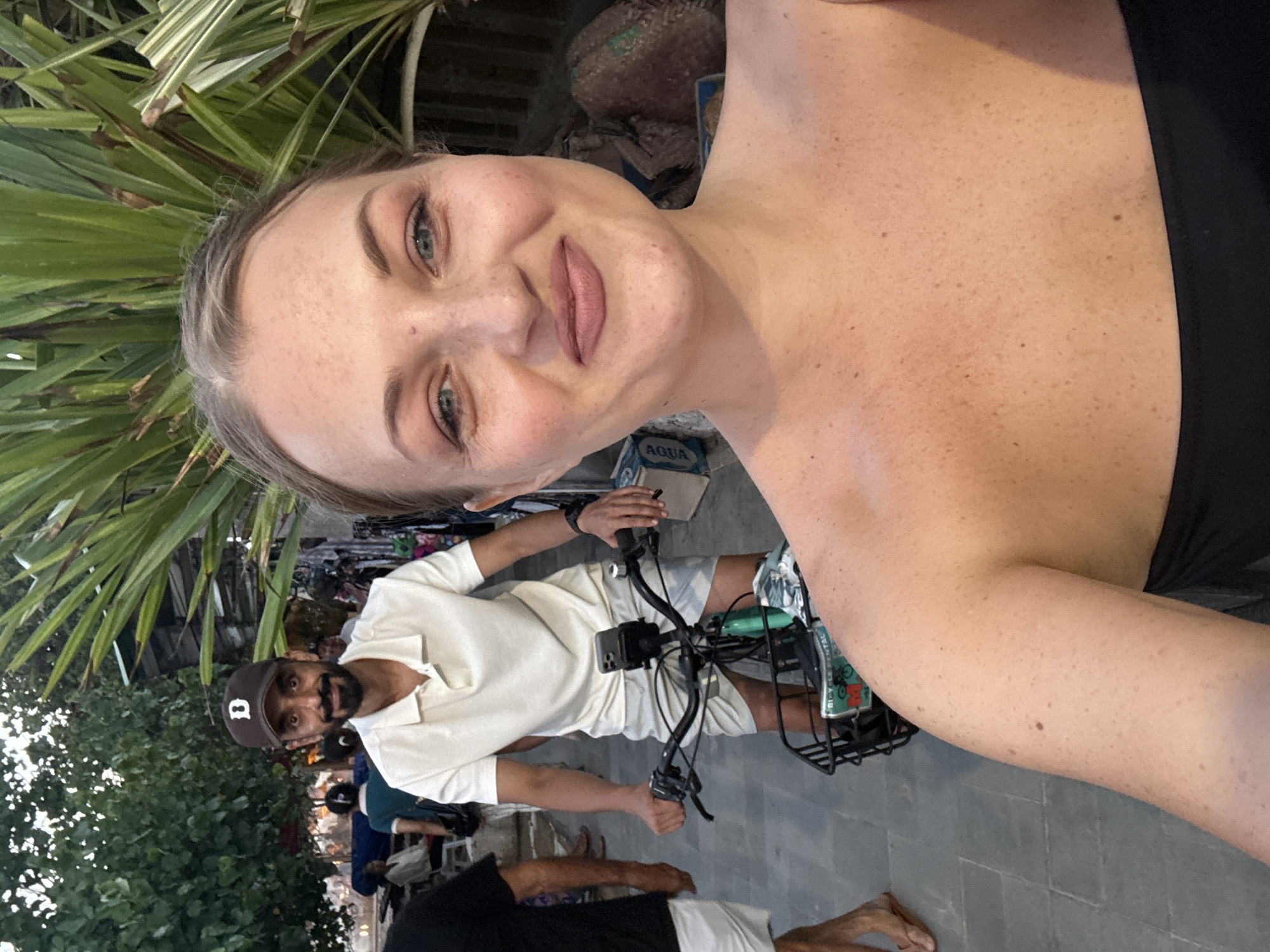 In Denpasar, Indonesia, a woman in the foreground takes a selfie. She has fair skin with freckles, blonde hair, and is wearing a black strapless top. In the background, a man wearing a white polo shirt, grey shorts, and a brown baseball cap with a "D" logo is riding a bicycle. The bicycle has a basket in the front. They are on a paved walkway, possibly a street, with lush green foliage behind them and other people in the distance. The lighting suggests it is either early morning or late afternoon.