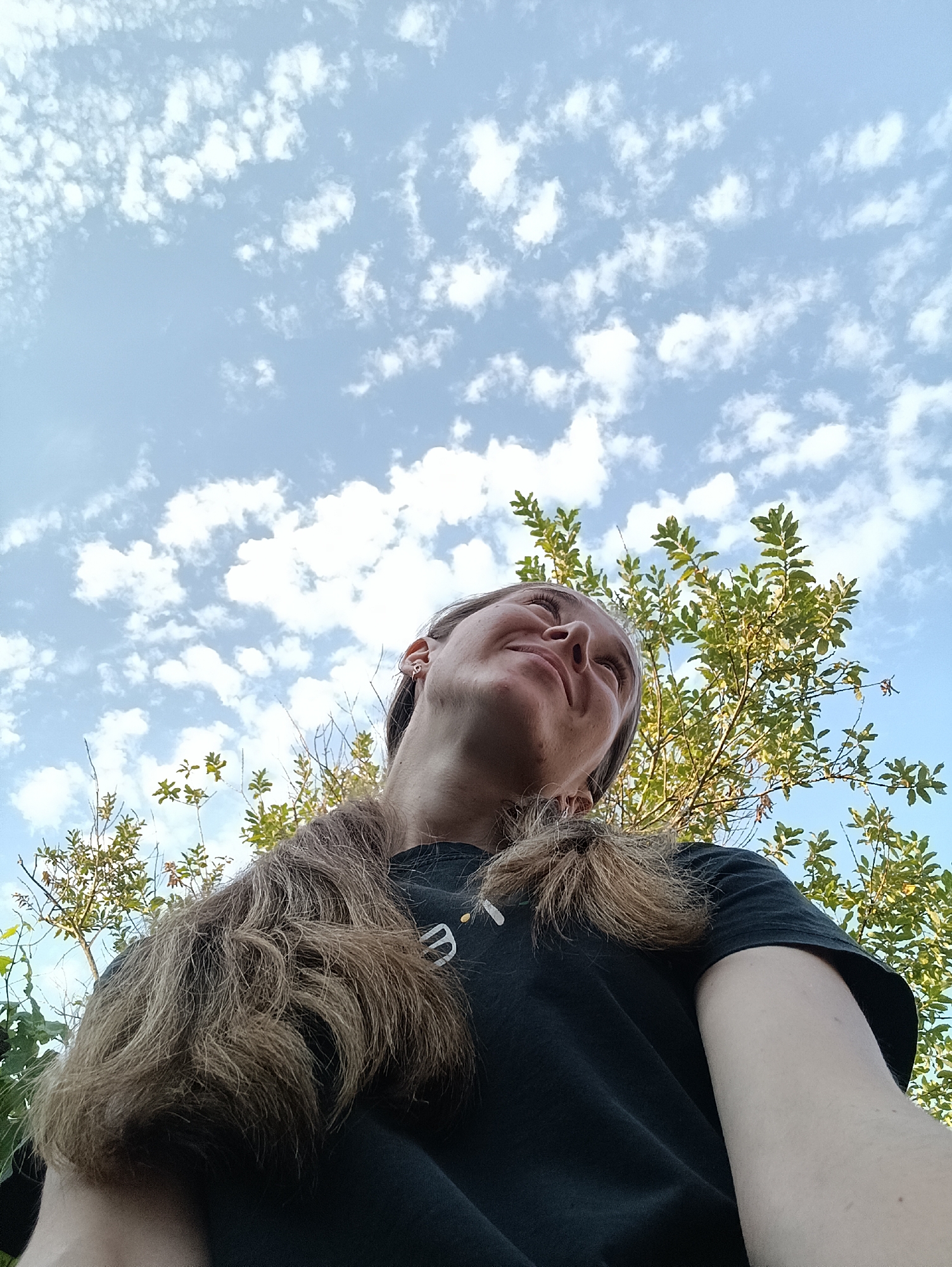 A low-angle outdoor shot shows a person with long, light brown hair looking up at a blue sky with scattered white clouds. The person is wearing a black t-shirt with a white logo. Green foliage from a tree is visible behind and to the right of the person. The scene appears to be in Vypasne, Ukraine.