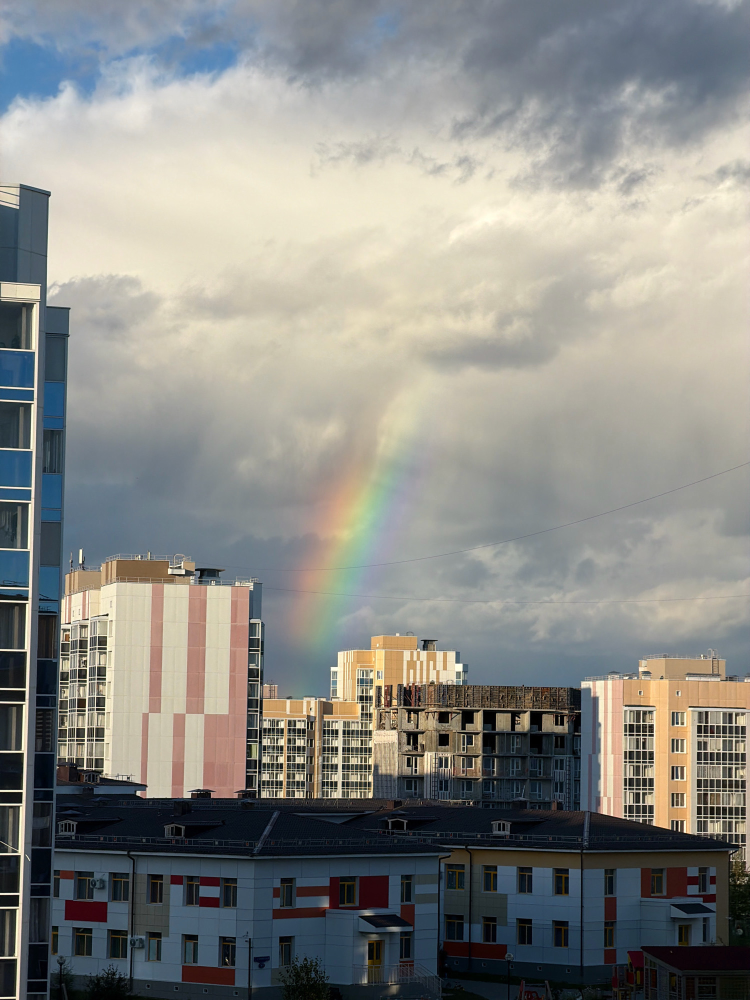 The image depicts a city scene in Tsiolkovsky, Russia, under a dramatic sky featuring a rainbow. In the foreground, several multi-story apartment buildings are visible, with varying architectural styles and colors. One building on the left is tall and slender with a blue and white facade. Adjacent to it is a building with vertical pink and white stripes. In the middle ground, a building under construction with visible concrete structures stands out. Further to the right, a pale yellow building with a grid of windows is present. In the foreground, a lower building with a colorful facade of white, red, and yellow sections, along with multiple windows, is also visible. The sky is filled with a mix of light and dark clouds, with the rainbow arcing through the upper portion of the image. Power lines are stretched across the sky in the middle distance. No people are visible in the frame.