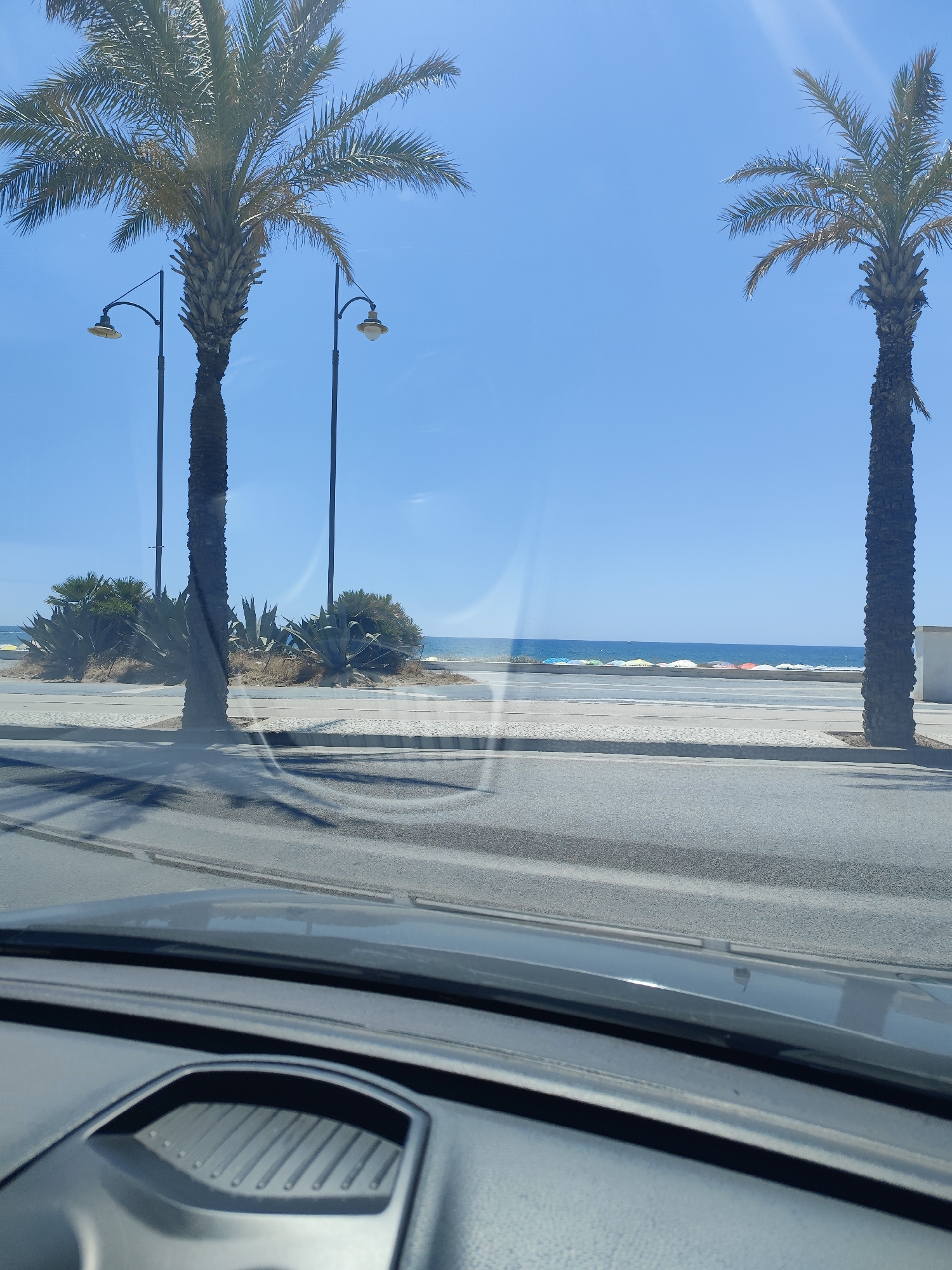 The image, taken from inside a vehicle, shows a sunny day at the beach in Mondragone, Italy. Two palm trees are visible, one in the left mid-ground and another in the right mid-ground. In between them, a row of beach umbrellas of various colors are scattered along the shoreline. The sea is calm and stretches towards the horizon, with a clear blue sky above. In the foreground, the dashboard and windshield of the car are visible, reflecting the outdoor scene. The road in front of the beach has tire marks.