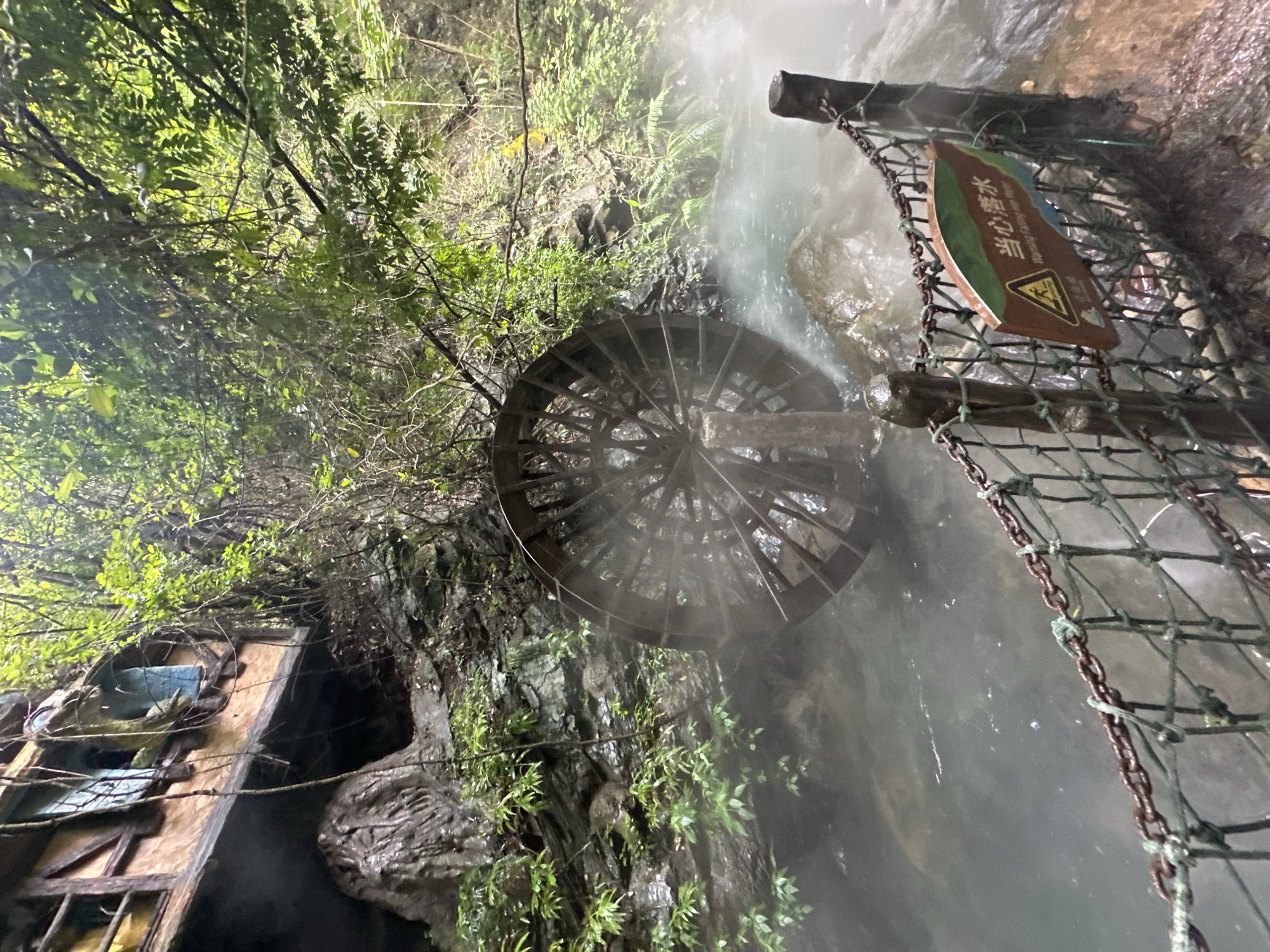 The image shows a water wheel in a stream, with a wooden structure built into a rocky cliff face behind it. The area is lush with vegetation, and a mist or steam rises from the water. A signpost with Chinese characters and an English warning about falling into the water is attached to a chain-link fence in the foreground. The water wheel appears to be made of wood and is partially submerged in the water, with its spokes visible. The wooden structure on the cliff appears to be an unusual building with windows and what looks like a balcony. This scene is located in the city of Qingyuan City, China, specifically in the area of the Huangteng Gorge, which is known for its hot springs and natural scenery. There are no people visible in the image.