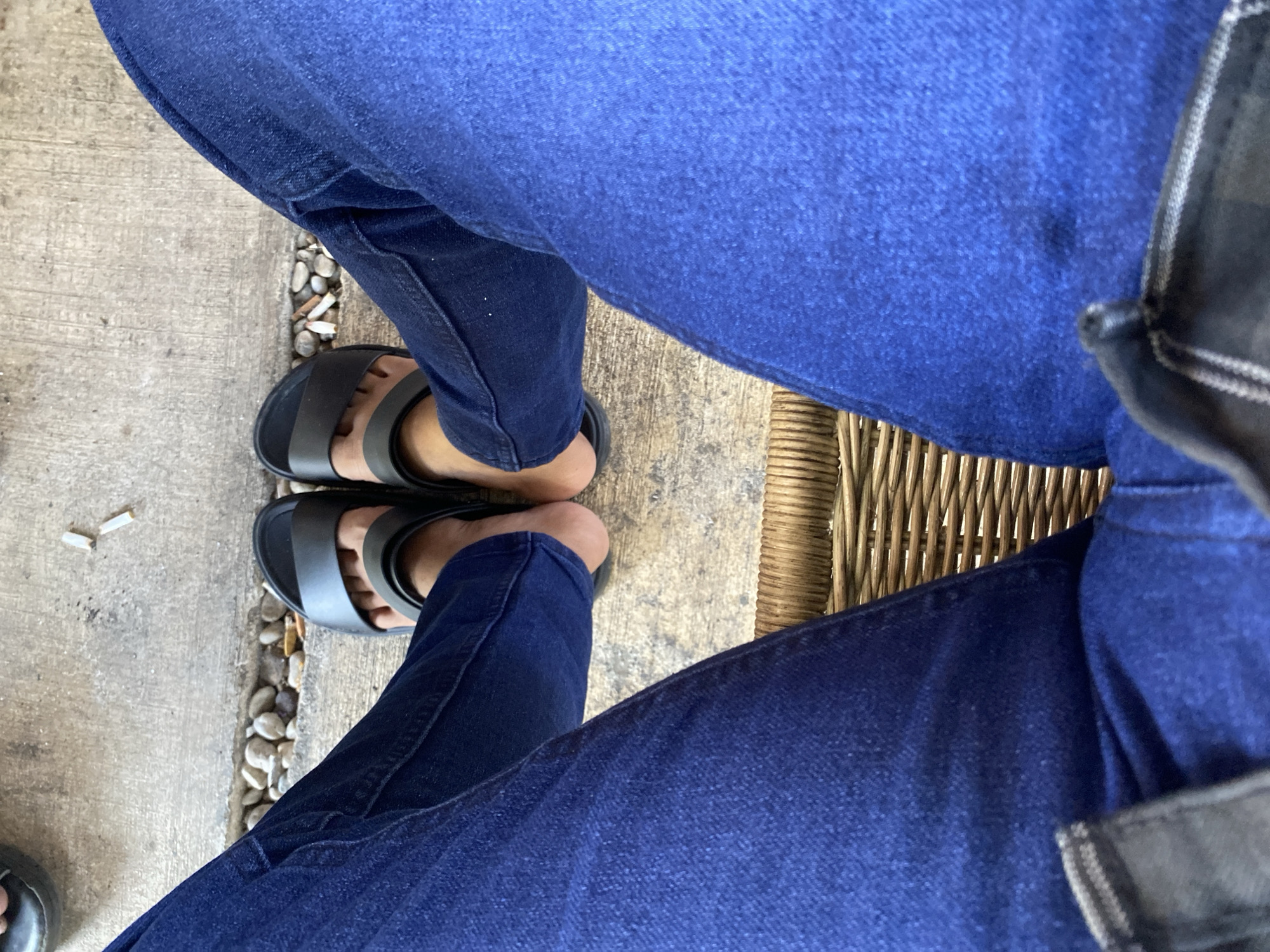 A first-person perspective shot shows a person sitting, with their legs crossed. The person is wearing dark blue denim jeans and black slide sandals. The background consists of a concrete walkway with small pebbles and two cigarette butts, and a woven cane chair. The image appears to have been taken outdoors in a location that might be found in Aluepiet, Indonesia, though no specific landmarks or distinctive city features are visible.