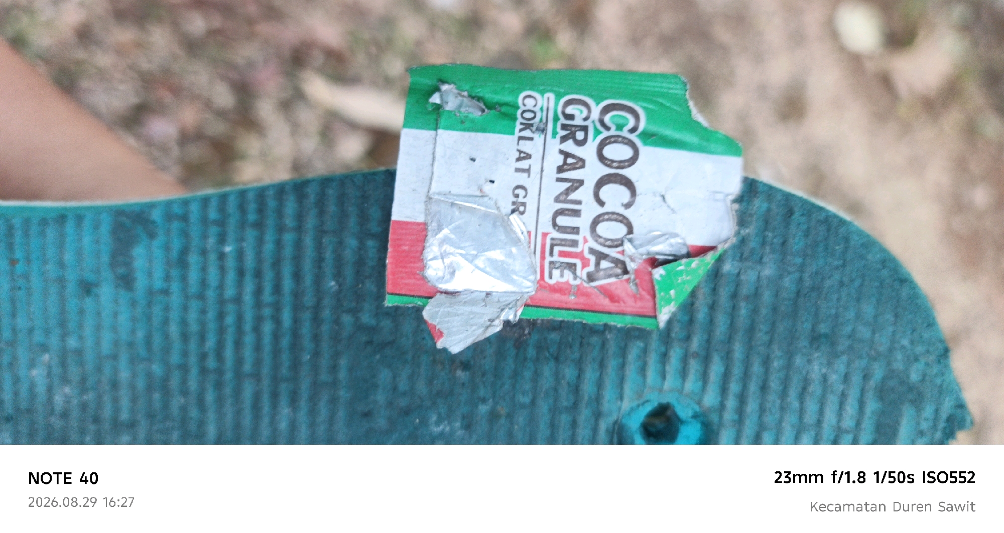 The image shows a close-up of a torn green and white wrapper for "COCOA GRANULE" with red lettering that reads "COKLAT GR". The wrapper is lying on a textured, teal-colored surface, possibly a flip-flop or a mat. In the background, out of focus, is a person's leg and a blurred, earthy-colored ground with some lighter colored objects. Text at the bottom of the image indicates the location as "Kecamatan Duren Sawit" in East Jakarta, Indonesia. The date and time are "2026.08.29 16:27" and the camera settings are "NOTE 40, 23mm f/1.8 1/50s ISO552".