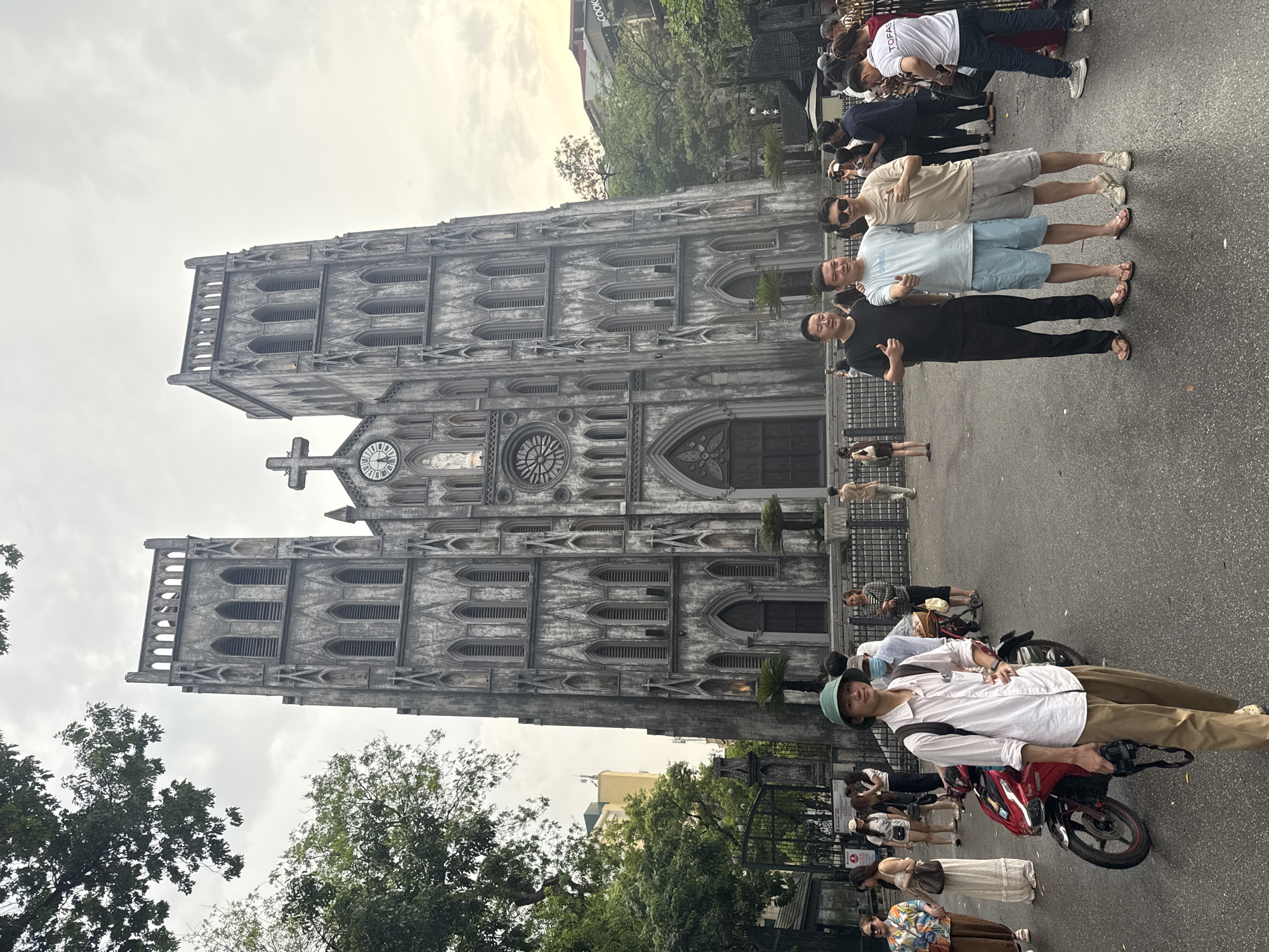 The image shows a wide shot of the St. Joseph's Cathedral in Hanoi, Vietnam. The cathedral is a neo-Gothic style church with two tall bell towers and a large clock face. In front of the cathedral, there are several people, including a group of three men posing for a photo in the foreground, a young man with a camera and a red motorcycle to his left, and other individuals and groups walking around. The ground is paved and there are trees and buildings visible in the background. The sky is overcast.