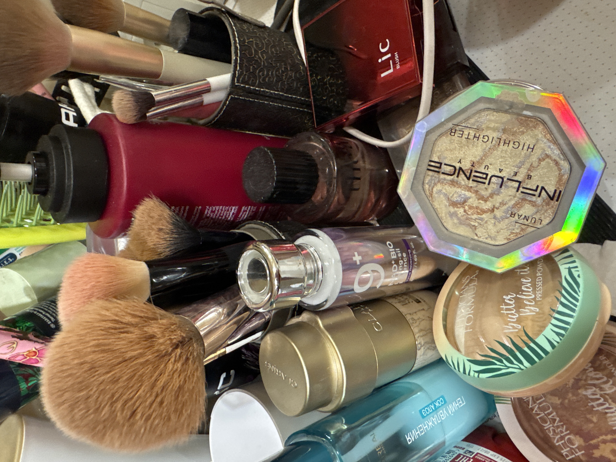 The image shows a close-up, high-angle shot of a collection of makeup and beauty products arranged on a surface. In the foreground, a hexagonal compact of "LUNAR INFLUENCE BEAUTY HIGHLIGHTER" is prominently displayed, with its iridescent packaging reflecting various colors. To its left, a round green-and-gold compact labeled "PHYSICIANS FORMULA Butter believe it PRESSED POWDER" is visible, with a partial view of another "PHYSICIANS FORMULA Butter Glow POWDER" compact below it.
Further into the image, a translucent blue bottle with Cyrillic lettering, likely a cosmetic product, is positioned to the left. Behind it, a tall gold-colored bottle with "CLARINS" embossed on its cap stands upright. Next to the gold bottle is a smaller, clear glass bottle with a silver cap and a label that reads "9+ BIO-SIL essence".
Dominating the upper portion of the frame are several makeup brushes with fluffy bristles in shades of brown and pink. They are housed in a dark, textured container. To the right of the brushes, a bright red bottle with a black pump dispenser is visible, along with a small dark bottle with a black cap, possibly perfume. A red compact labeled "Lic BLUSH" is also present in the right-middle section. The background is cluttered with various other items and appears to be an indoor setting.