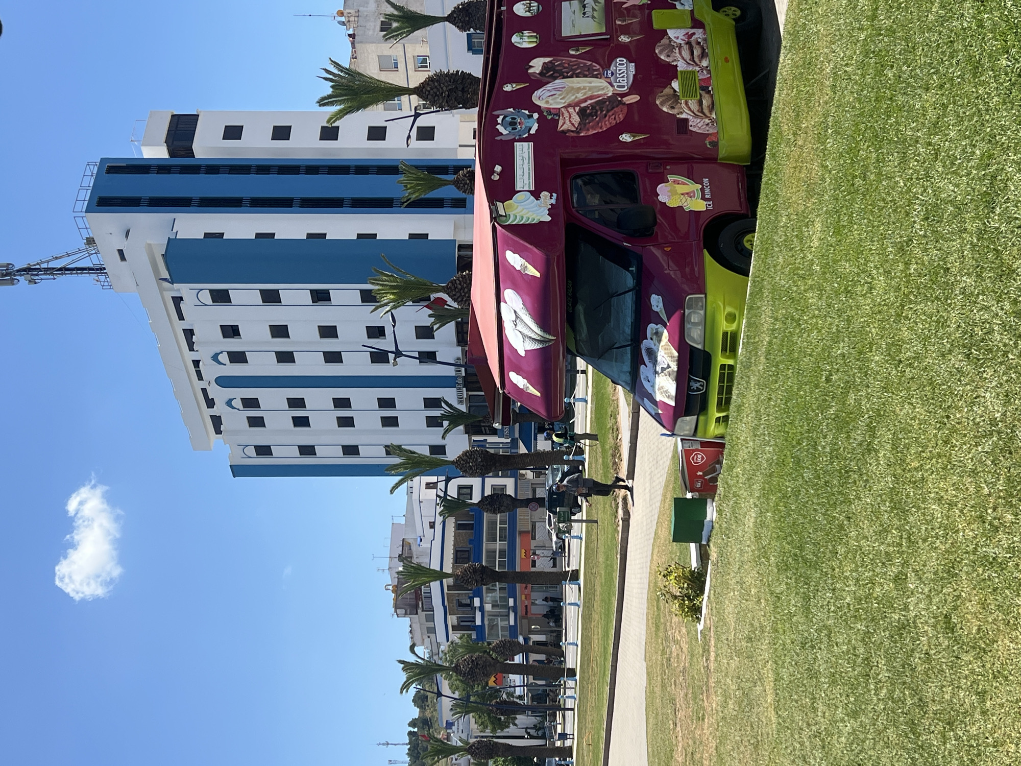 The image shows a street scene in M'Diq, Morocco. A burgundy and green ice cream truck with cartoon graphics is parked next to a green grassy area. Behind the truck is a sidewalk lined with palm trees. On the other side of the sidewalk are a series of buildings, predominantly white with blue accents. The sky is clear blue with a small white cloud in the upper left corner. People are walking along the sidewalk.