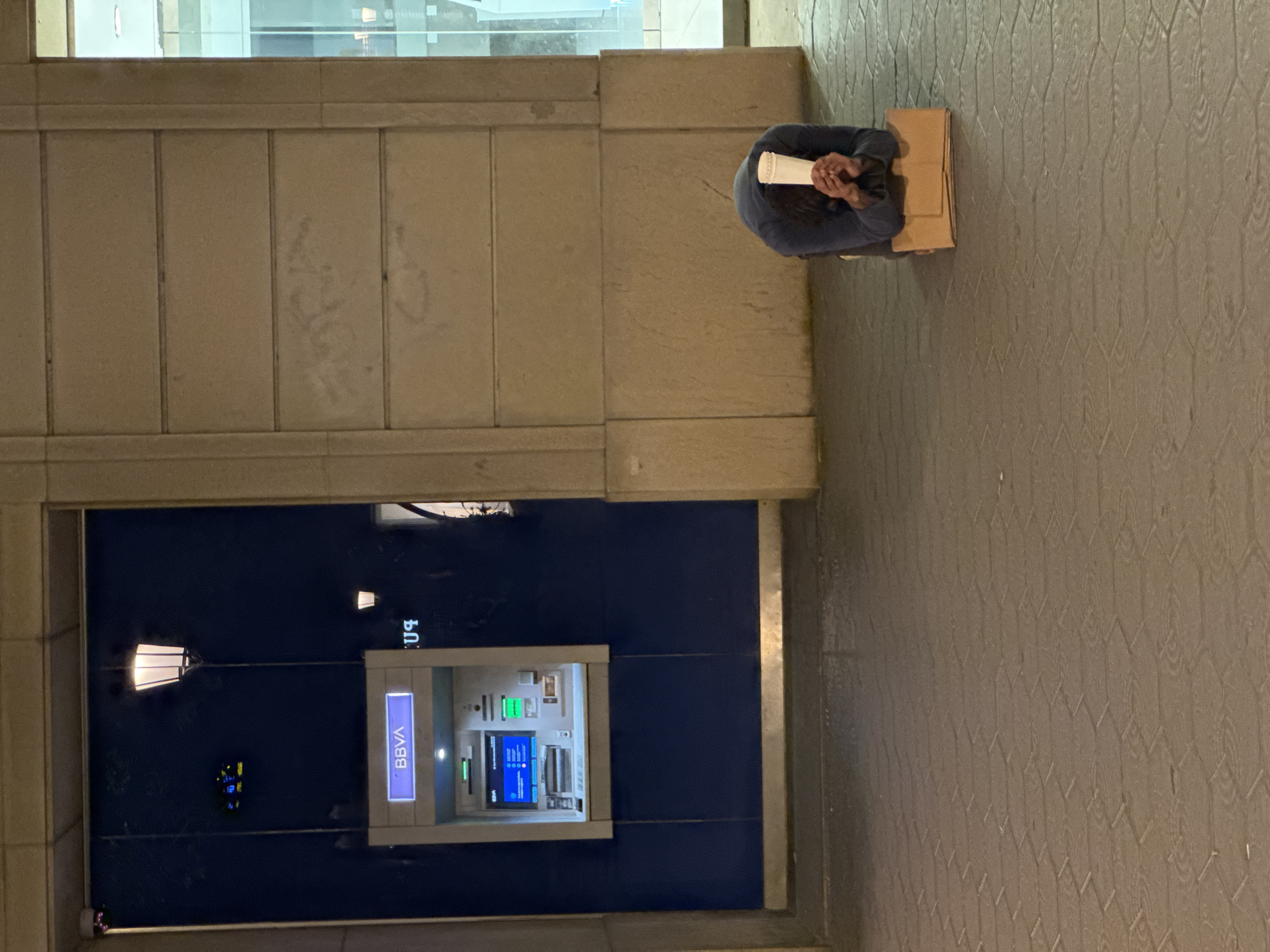A woman is sitting on the ground, on top of a cardboard box, in front of a BBVA bank ATM in Barcelona, Spain. She is dressed in a dark-colored long-sleeved shirt and is holding a white, cylindrical object in her hands. The ATM is illuminated and displays a screen with blue and green elements. A lit streetlamp is positioned to the left of the ATM. The scene is set against a building facade constructed of concrete blocks and tiled ground.