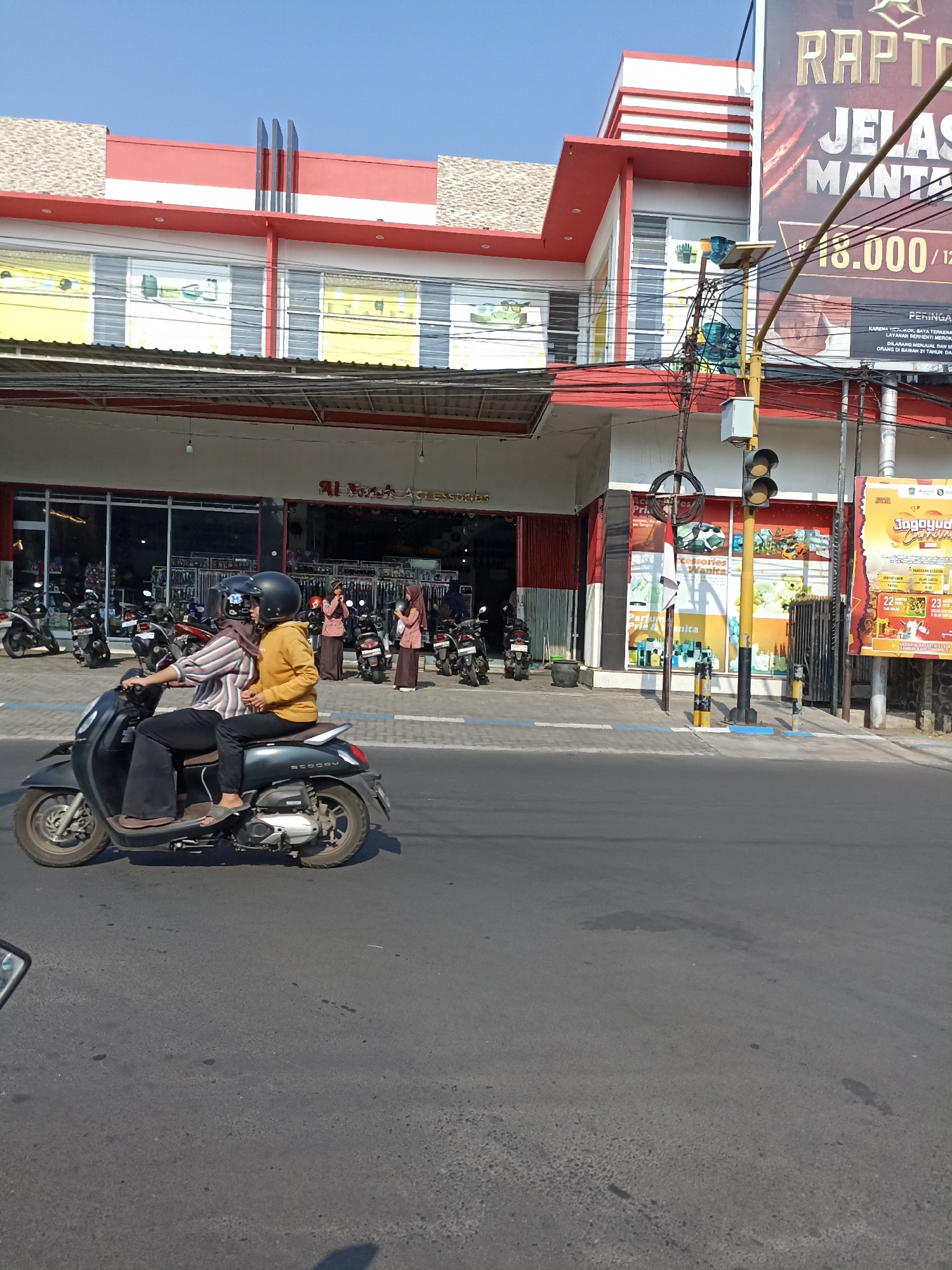 A street scene in Jogoyudan, Indonesia, under a clear blue sky, captures a moment on a sunny day. The foreground features a dark gray scooter with two occupants, a driver and a passenger, both wearing helmets. The driver is in a striped long-sleeved shirt and dark pants, while the passenger is wearing a yellow hooded jacket and dark pants. The scooter is moving from left to right.

Behind the scooter, a building with a red and white facade is visible, featuring large glass windows that reveal displays of merchandise. The building appears to be a store, possibly selling accessories, indicated by signage that includes "Aksesoris" and "Parfum Pria & Wanita". Several motorcycles are parked in front of the store, and three women in headscarves and long dresses stand near the entrance.

A yellow traffic light pole with a signal is positioned to the right of the building. A large billboard with text and images, likely advertising a product or event, is mounted on the side of another building to the right. The street is paved, with some dark patches suggesting water or oil stains. The overall environment suggests a commercial area in an Indonesian city.