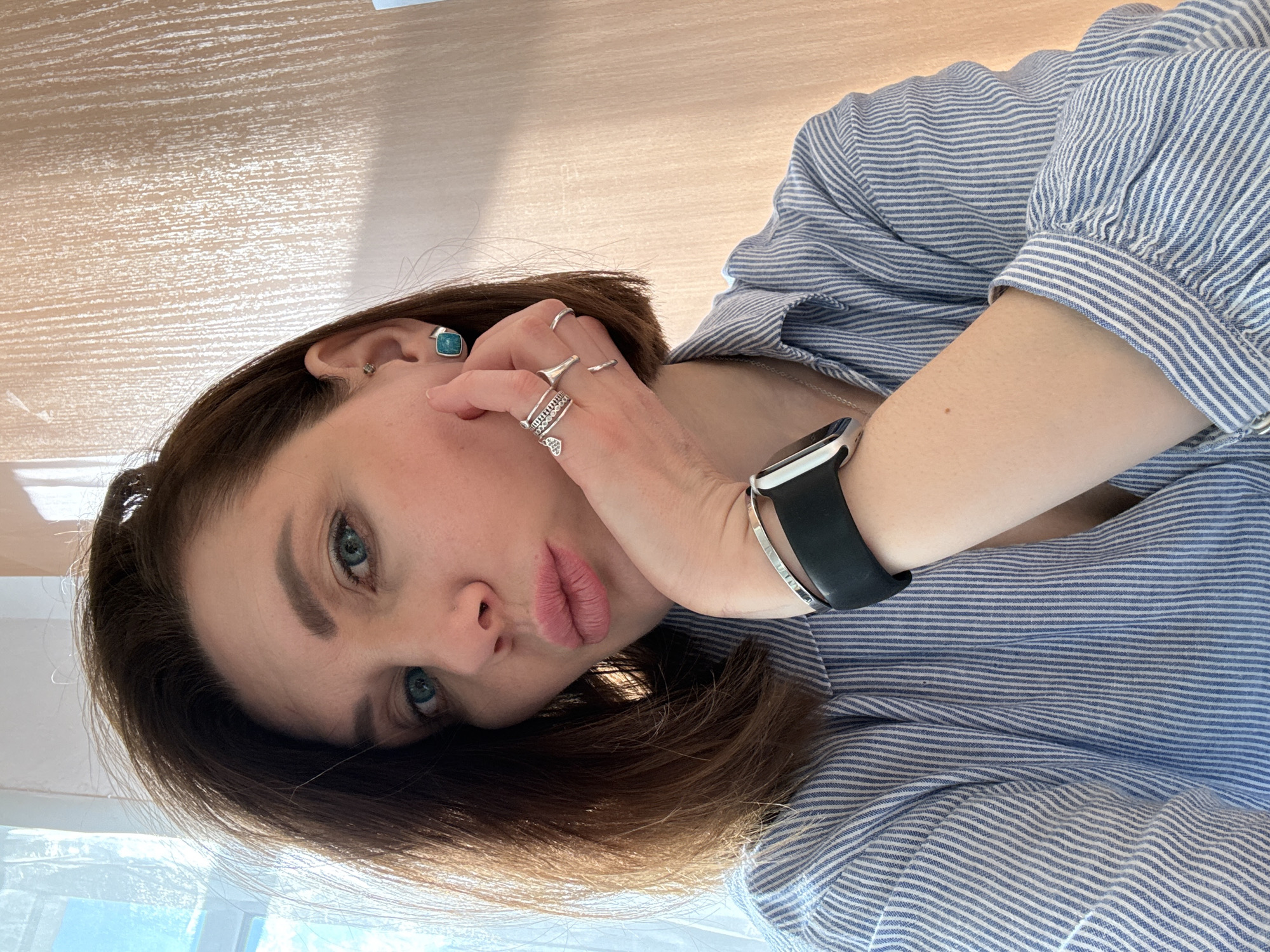 The image shows a woman with brown hair and blue eyes looking at the camera. She is wearing a blue and white striped shirt. She has several rings on her fingers and a smartwatch on her wrist. She has turquoise stud earrings in her ears. The background appears to be a wooden surface. The location is unknown and no clues are in the picture to assume the location is in the city of Chaikovsky, Russia.
