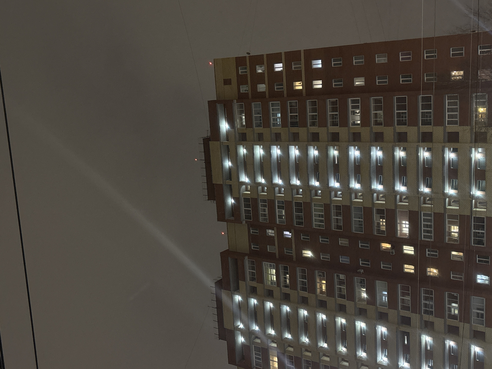 The image captures a tall building in Perm, Russia, during the night. The building has a patterned facade, with alternating sections of dark brick and lighter-colored material. Windows are visible throughout the building, some illuminated from within. Vertical light fixtures are affixed to the building's exterior, casting light downwards. Wires stretch across the upper portion of the building and into the dark, overcast sky.