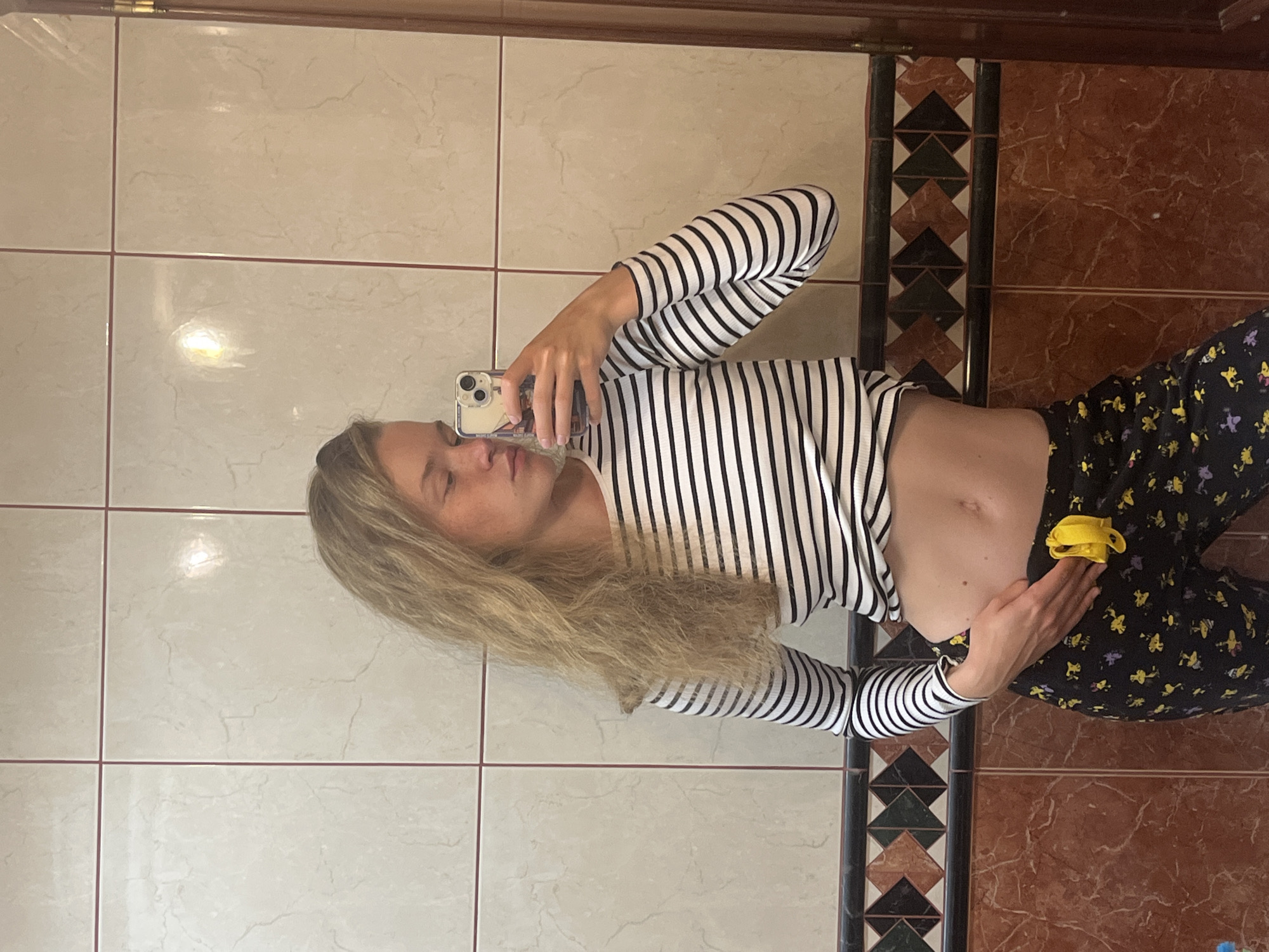 A young woman with blonde hair captures a mirror selfie in what appears to be an indoor setting, possibly a bathroom or hallway, located in Budva, Montenegro. She is wearing a black and white striped long-sleeve crop top, revealing her midriff, and black patterned pants adorned with yellow and purple motifs, resembling pajama bottoms. On her right wrist, she wears a yellow hair tie or scrunchie.
The woman is holding a smartphone, which features a triple-lens camera, with her right hand to take the photo, and her gaze is directed towards the phone screen. Her left hand is gently placed on her stomach.
The background consists of tiled walls. To her left, the tiles are light beige with thin reddish grout lines. To her right, the tiles are a darker reddish-brown with a marble-like pattern. A decorative vertical border, featuring geometric patterns in black, white, and brown, separates these two tile sections. The lighting in the room is soft and even. There is no visible text in the image.