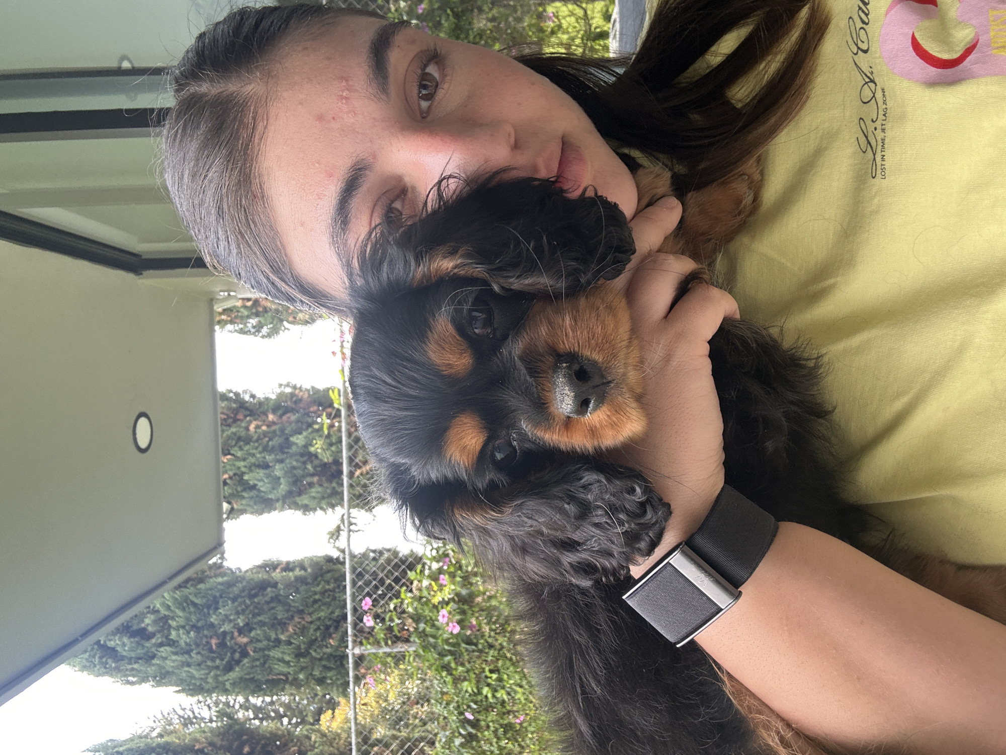 A woman with brown hair holds a black and brown Cocker Spaniel puppy close to her face. She has brown eyes and light skin. She wears a yellow t-shirt. A black smartwatch is visible on her left wrist. The background shows greenery, a fence, and a building. The location is Puebla, Mexico.
