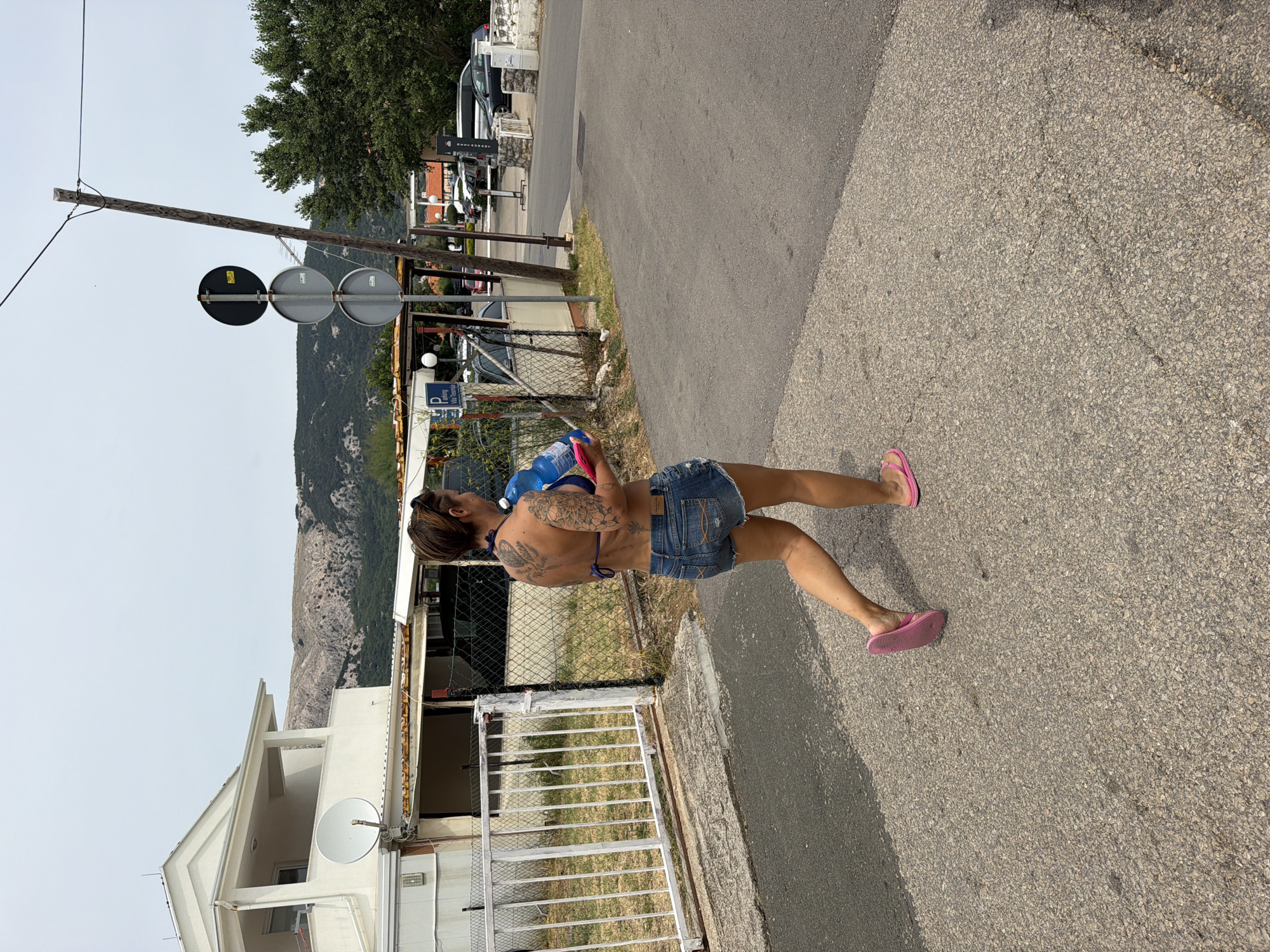 A woman with tattoos on her back walks on a paved road in Općina Baška, Croatia. She is wearing a blue bikini top, denim shorts, and pink flip-flops. She is carrying a large blue water jug and a pink object in her arms. To her left is a white fence and a building with a satellite dish. In the background, there are trees, cars, and a hilly landscape under a cloudy sky. A signpost with three circular signs is visible to her right.