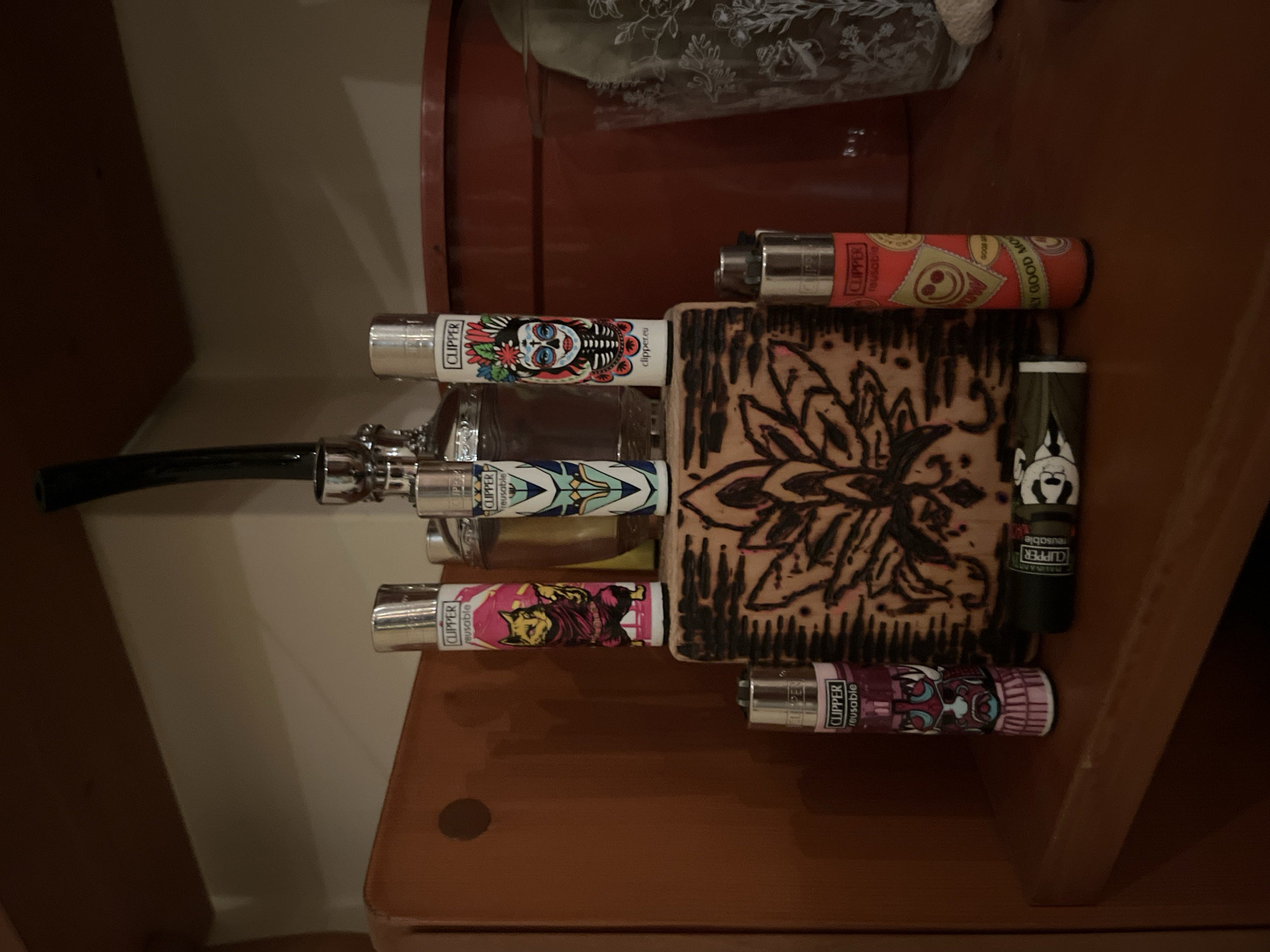 The image displays a collection of seven Clipper lighters arranged on a wooden surface within an indoor setting. A central, rectangular wooden block features a dark, stylized floral or lotus design etched into its surface. Three lighters stand upright on this block: one pink with a yellow cat figure, one white with a blue and gold geometric pattern, and one white with a colorful "Day of the Dead" sugar skull woman. Two additional lighters stand on the wooden surface in front of the block: one dark pink with a mask-like design, and one orange with yellow smiley faces and "GOOD MOOD" text. A seventh black lighter lies horizontally in front of the block, displaying a white skull-like face. All visible lighters are marked "CLIPPER reusable". In the background, a clear glass bottle with a silver dispenser and a dark, curved hose is visible. To the right, a large reddish-brown pot and a clear glass with a frosted floral pattern sit on the surface, adjacent to a small white textile item. The environment consists of wooden shelves or furniture in warm brown tones and a light-colored wall, illuminated by warm, dim lighting. The image provides no discernible information to suggest a specific location in Varna, Bulgaria.