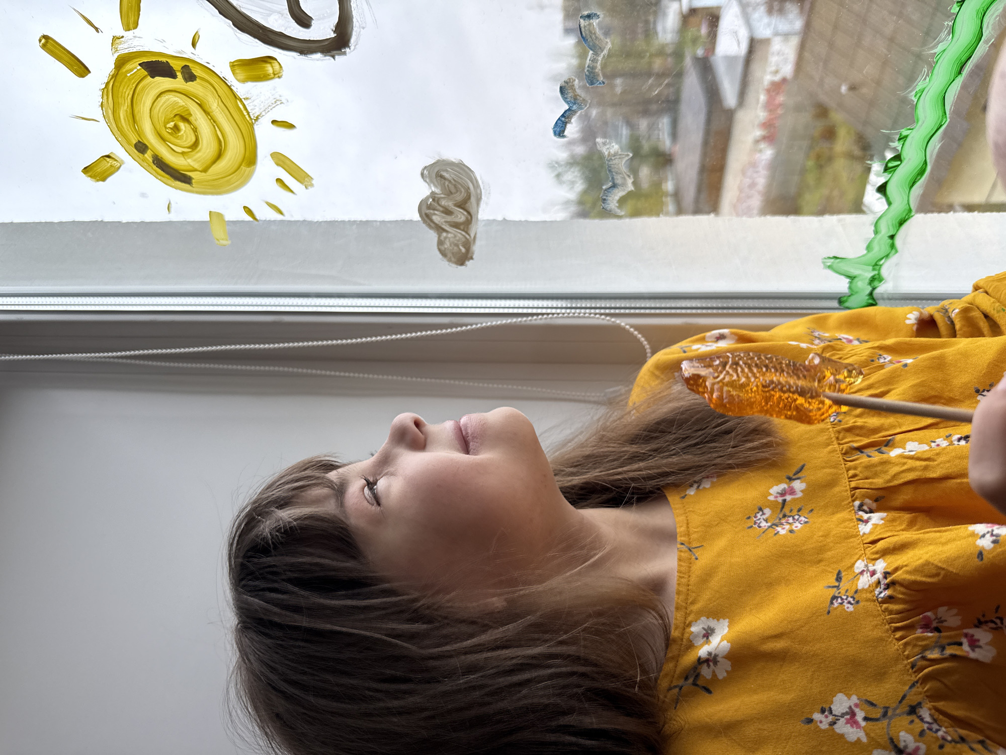 A young girl is depicted in a side profile, positioned horizontally in the frame, looking upwards and to the left with a serene expression, her eyes partially closed. She has long, dark brown hair and is wearing a mustard yellow dress adorned with a white and pink floral pattern. In her right hand, she holds an amber-colored, translucent lollipop on a stick, which appears to be either a sweet treat or an unconventional painting tool.
The girl is positioned against a window, which is decorated with colorful, childlike paintings. On the upper left, a vibrant yellow sun with radiating brushstrokes dominates the glass. To its right, a dark brown swirling mark and a white or beige cloud-like smudge are visible. Further to the right, blue wavy lines, possibly representing birds or abstract elements, are painted. Along the far right edge of the window, a bright green wavy line stretches down, resembling a plant stem or abstract design.
Through the unpainted sections of the window, a blurred outdoor scene is visible, showing hints of buildings and foliage, suggesting an urban or suburban setting. The lighting is soft and diffused, characteristic of an overcast day. A white window sill and a pull cord for blinds are also visible at the bottom of the window. The overall image captures a moment of creative play and peaceful concentration, possibly engaging in temporary window art. There is no visible text in the image.