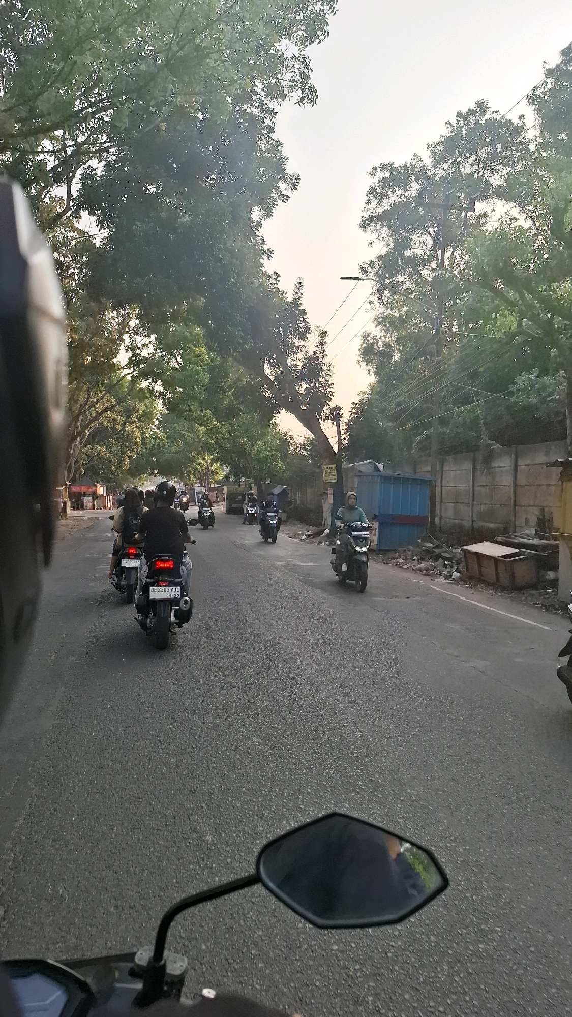 The image shows a view from a motorcycle in the city of Bandar Lampung, Indonesia, on a street lined with trees. The sky is overcast, suggesting it might be morning or late afternoon. Several motorcycles are visible on the road, indicating traffic. One rider in the foreground, wearing a black helmet, is looking ahead. In the middle ground, other motorcycles with riders are proceeding down the street. On the right side of the road, there is a concrete wall and some debris. A person in a hijab is riding a scooter on the right side of the road. The overall impression is a typical urban street scene in Bandar Lampung.