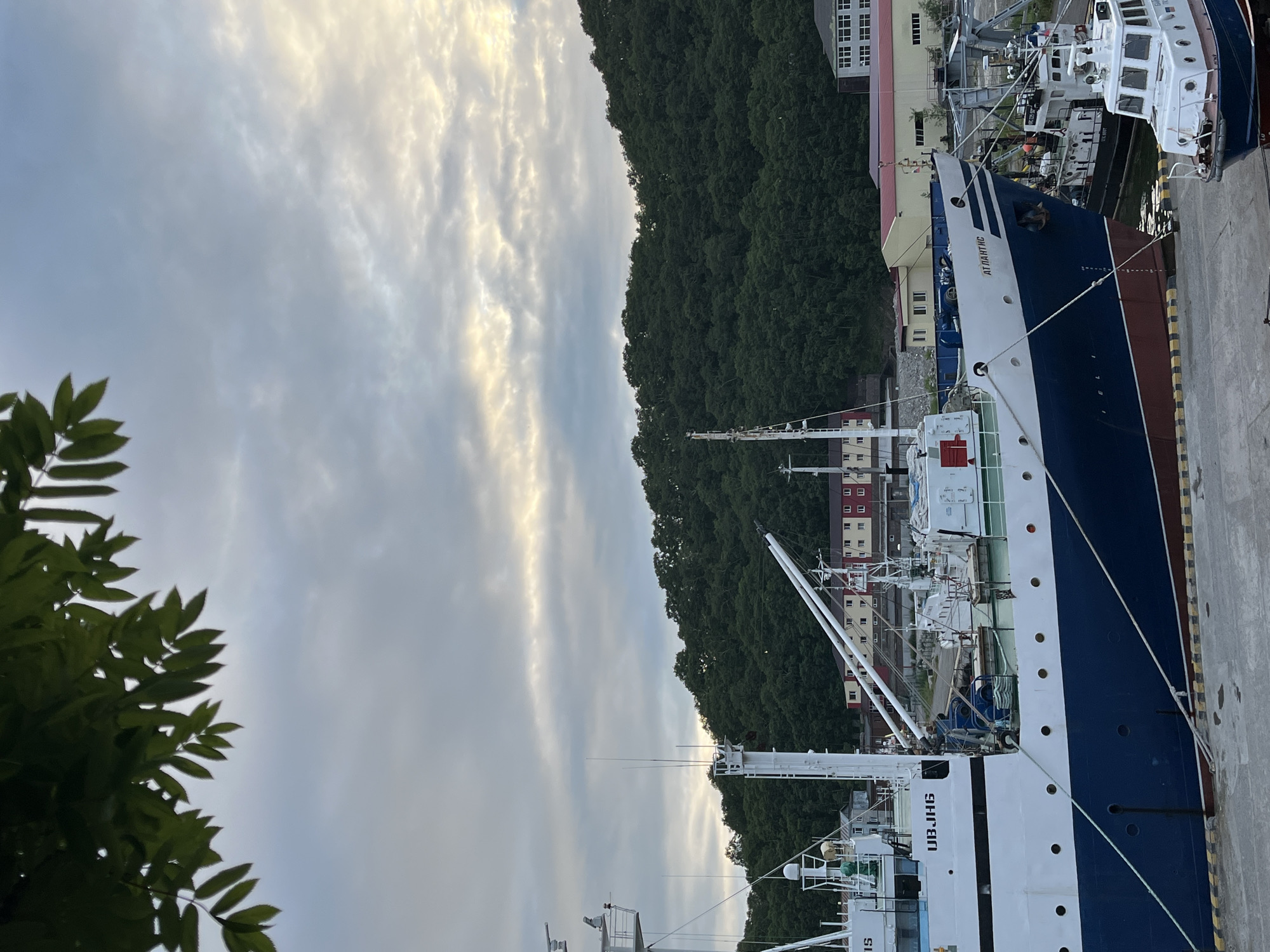 A long shot captures a view of a harbor in Petropavlovsk-Kamchatsky, Russia, with a large blue and white ship docked in the foreground. The ship has the Cyrillic lettering "АТЛАНТ ПС" on its bow and "UBJHG" on its side. In the background, a lush green, tree-covered hill rises steeply. Several buildings, including what appears to be a red-roofed structure and a multi-story building with white and tan facades, are situated at the base of the hill. The sky is cloudy, with streaks of sunlight breaking through. In the lower right corner, a smaller white boat with blue trim is docked. The overall scene suggests a working harbor under a partly cloudy sky.