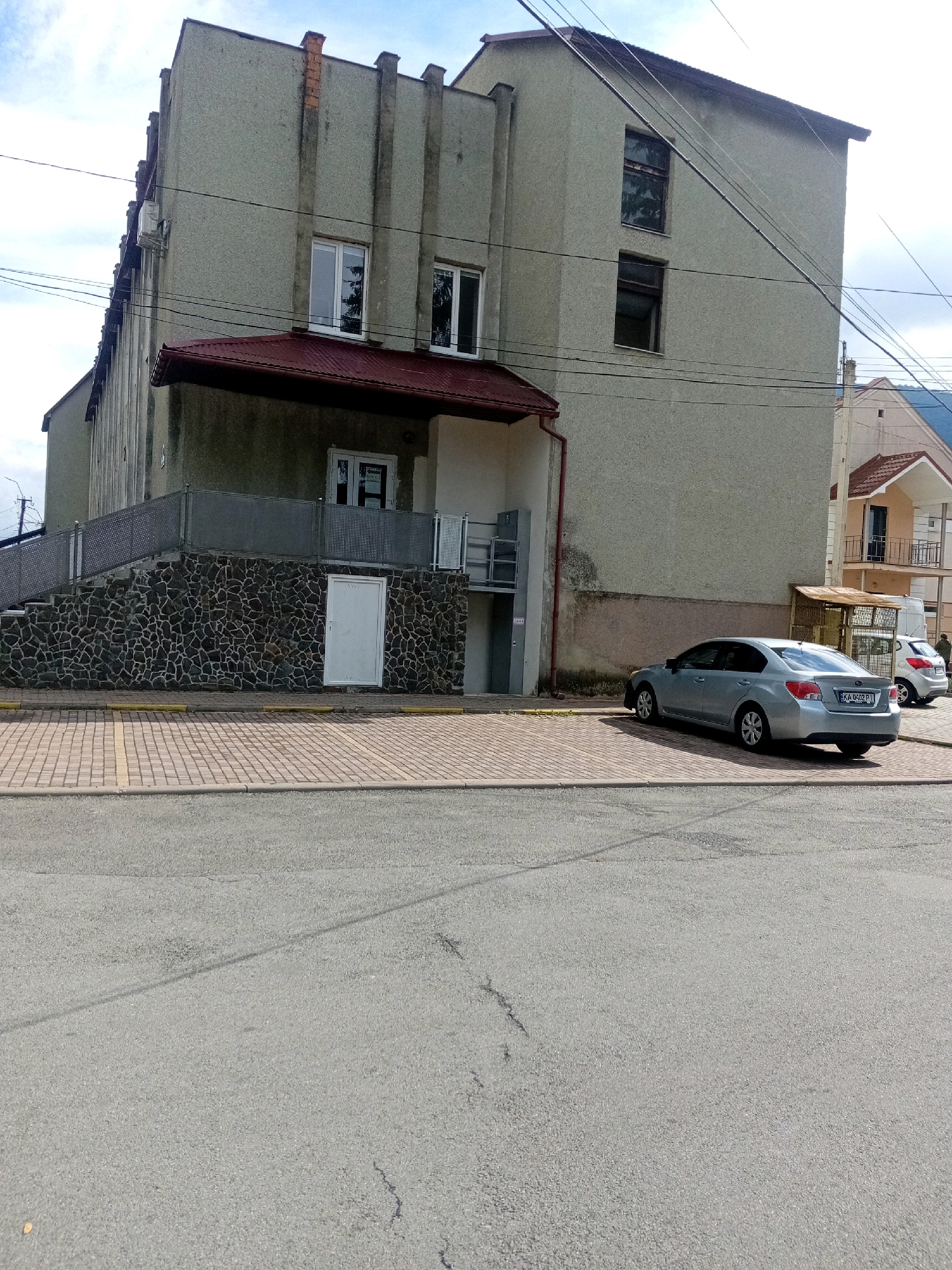 A medium shot captures a light blue sedan parked on a cobblestone lot in front of a light gray, multi-story building in Tiachiv, Ukraine. The building features a dark red, sloped roof over a section of its entrance, which is adorned with a stone facade. A white door is visible on the stone section. The sedan's license plate, visible on the rear, reads "KA 0402 PI". To the right of the sedan, a white hatchback is partially visible. The asphalt street runs in the foreground. Power lines crisscross the sky above the scene. The sky is partly cloudy.