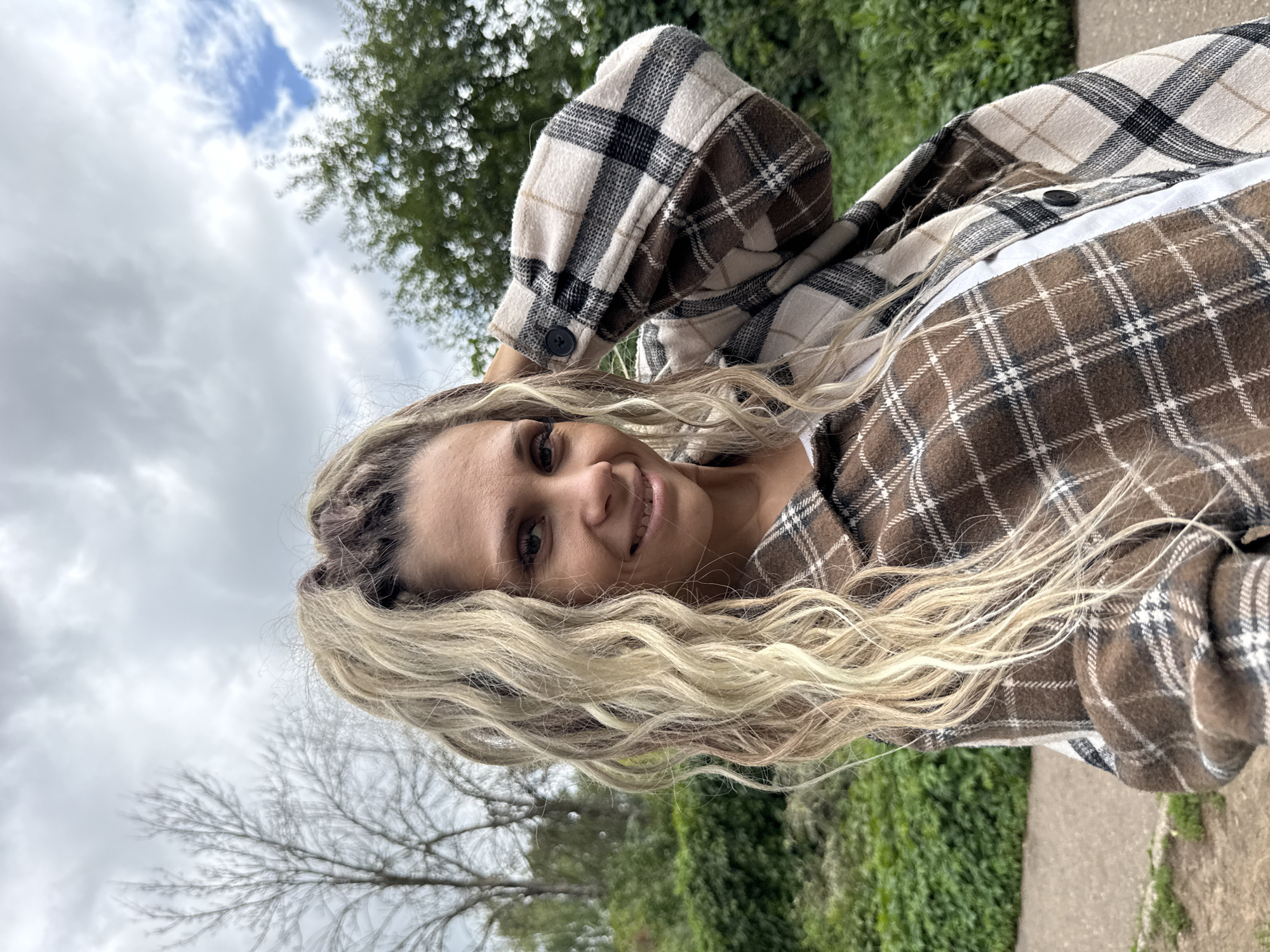 A waist-up, medium shot captures a woman with blonde, wavy dreadlocks, styled into a high ponytail, posing outdoors. She wears a brown, cream, and black plaid shirt jacket over a white garment. Her right arm is bent, with her hand resting behind her head, and she smiles at the camera. The background features lush green foliage and trees under a cloudy sky with patches of blue. A paved path is visible in the lower left quadrant of the image.