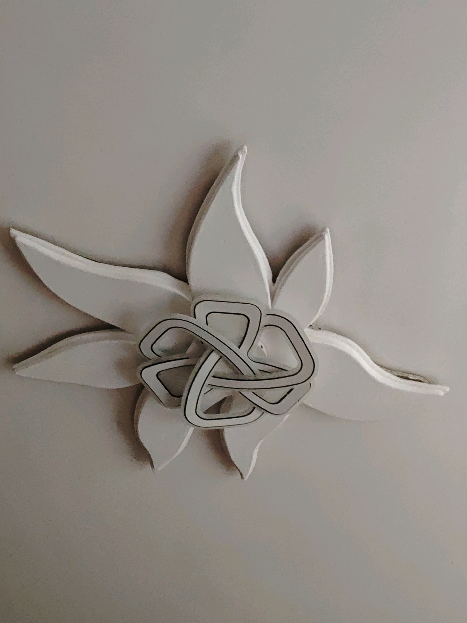 The image shows a close-up, overhead view of a white decorative element, resembling a stylized flower, mounted on a light-colored, textured surface, likely a ceiling. The flower has multiple pointed petals radiating outwards. In the center of the flower is an intricate, interwoven geometric pattern made of white material with thin black outlines. The lighting creates subtle shadows, highlighting the three-dimensional nature of the object. There are no people, animals, or any other identifiable objects or actions visible. The location in Ninotsminda, Georgia, is not discernible from the visual information provided in the image.