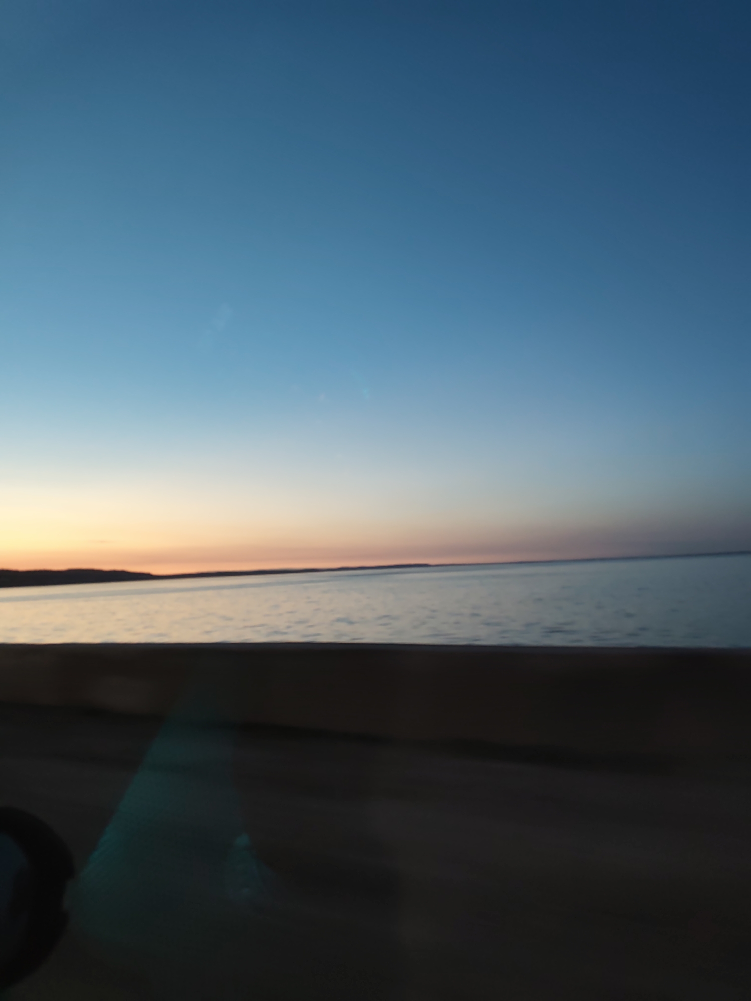 The image depicts a sunset over a body of water, likely a lake or a wide river. The sky transitions from a deep blue at the top to lighter blue and then to orange near the horizon. A dark shoreline can be seen in the distance, possibly the opposite bank of the river or lake. In the foreground, there is a blurred, dark surface, possibly the dashboard of a moving vehicle, with a hint of a reflection on the windshield. The photo appears to have been taken from inside a car. The location is said to be Chaikovsky, Russia. The image does not show any people, interactions, or precise details of the environment beyond the body of water and shoreline.