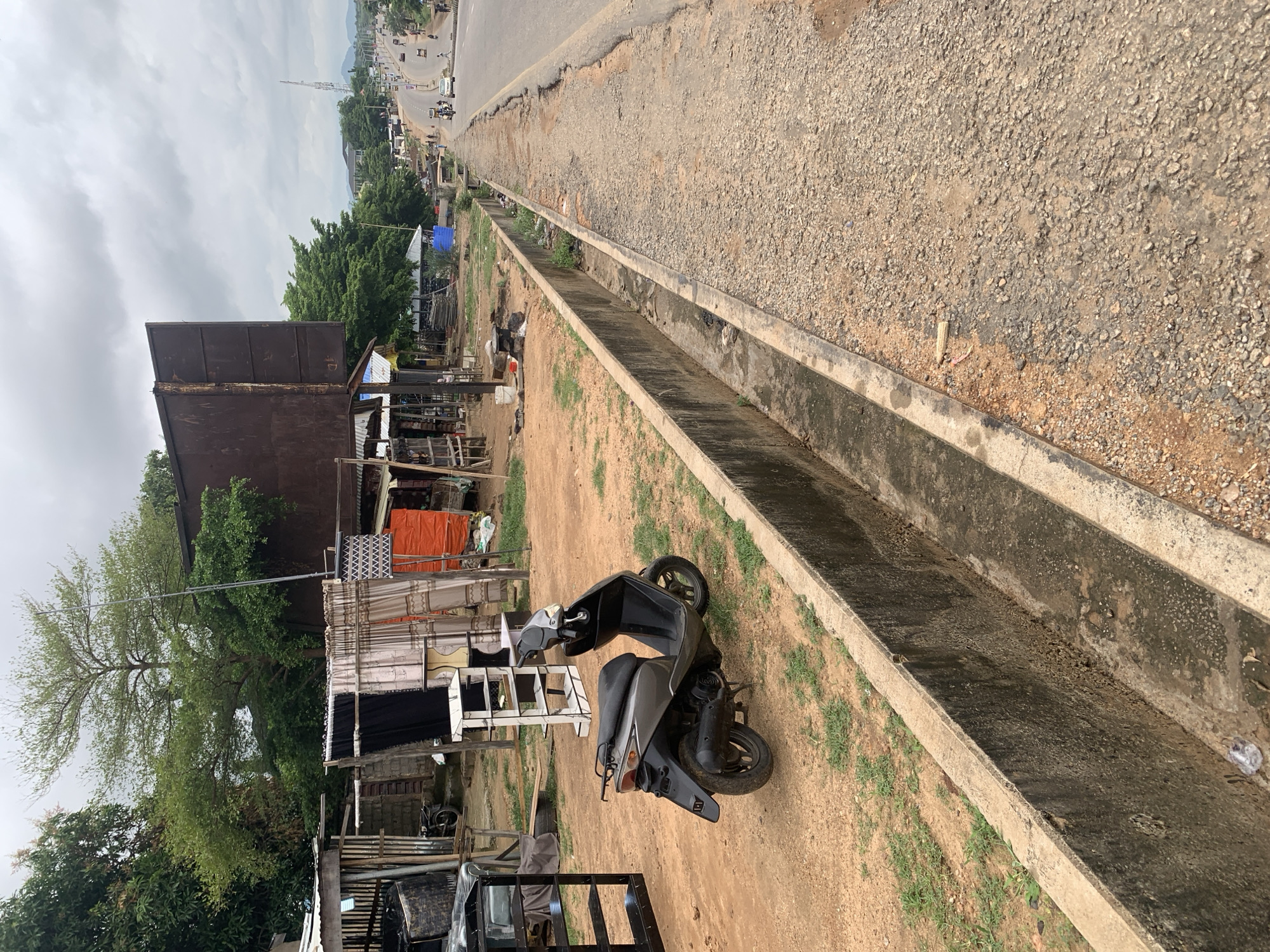 The image shows a street scene in Adankolo, Nigeria, with a grey scooter parked on the side of a dirt road next to a concrete drainage ditch. Behind the scooter, a makeshift market stall is visible with various items displayed, including fabrics and what appear to be shelving units. The road stretches into the distance, lined with buildings and trees, and traffic is visible, including cars and motorcycles. The sky is overcast.