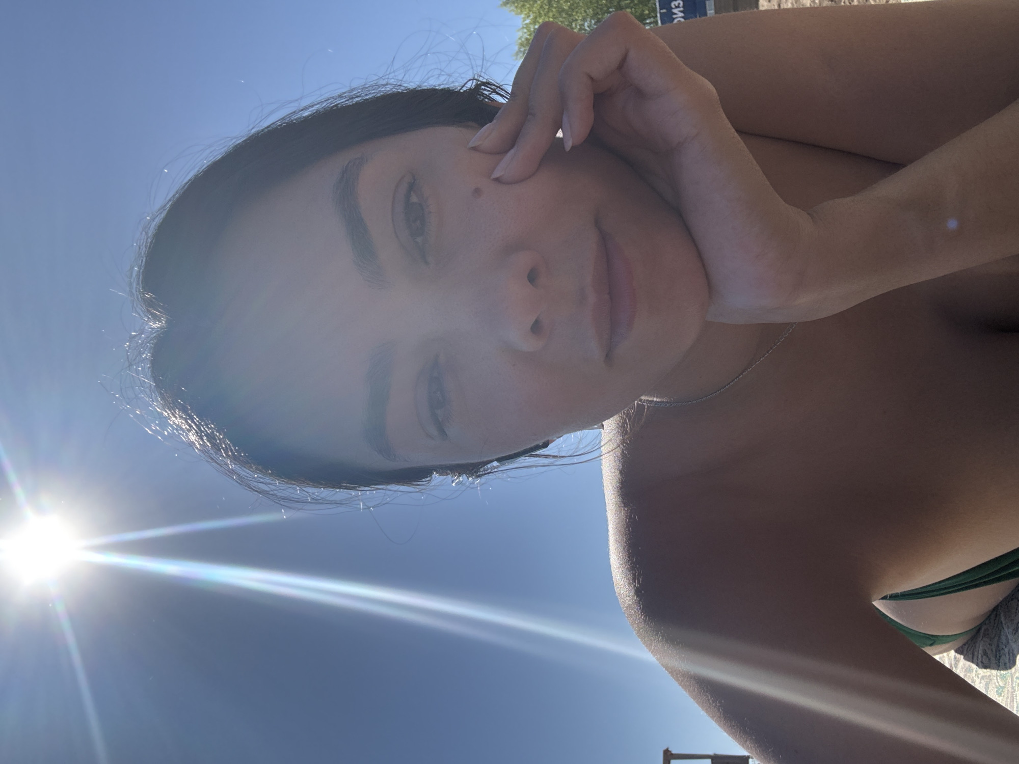 A selfie taken outdoors on a sunny day. A woman with dark hair is posing with her hand near her face. She is wearing a green bikini top. The background is a clear blue sky, with bright sunlight causing lens flare. Based on the text visible in the background (cropped out), the photo was likely taken in the city of Chaikovsky, Russia.