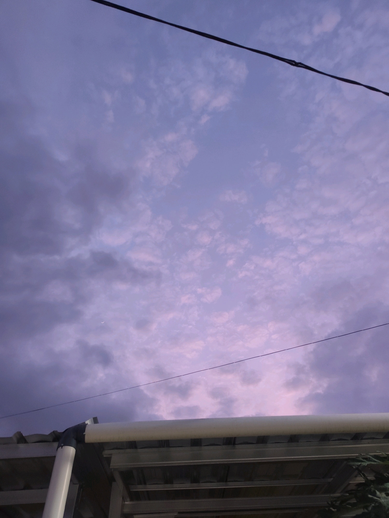 The image depicts a dusk sky over the city of Bambanglipuro, Indonesia. The sky is filled with scattered clouds, displaying shades of purple, blue, and pink. A thin crescent moon is visible in the upper left quadrant. In the foreground, a portion of a building's roof structure is visible, including white and grey pipes, and metal framing. A black wire stretches diagonally across the upper portion of the image. No people are present in the scene.