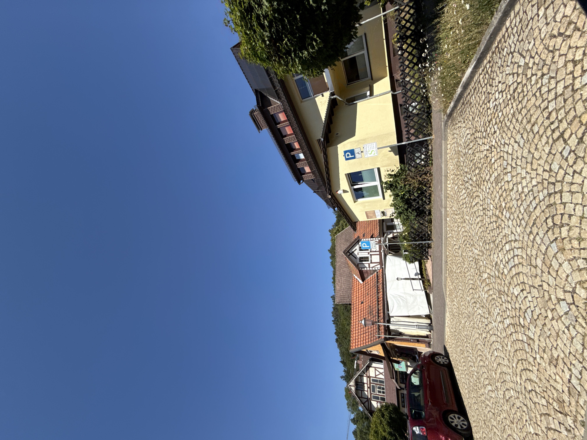 The image captures a sunlit street scene in Bad Sachsa, Germany. In the foreground, a cobblestone surface is visible, forming a parking area or plaza. To the left, a red hatchback car is parked. Behind the car, a row of buildings with traditional German architecture lines the street. These buildings feature pitched roofs, some with red tiles, and elements of half-timbered construction. One building is painted yellow and has solar panels on its roof. Parking signs are visible in front of the buildings. In the background, a dense green forest rises, meeting a clear blue sky. The overall atmosphere is one of a quiet, sunny day in a German town.