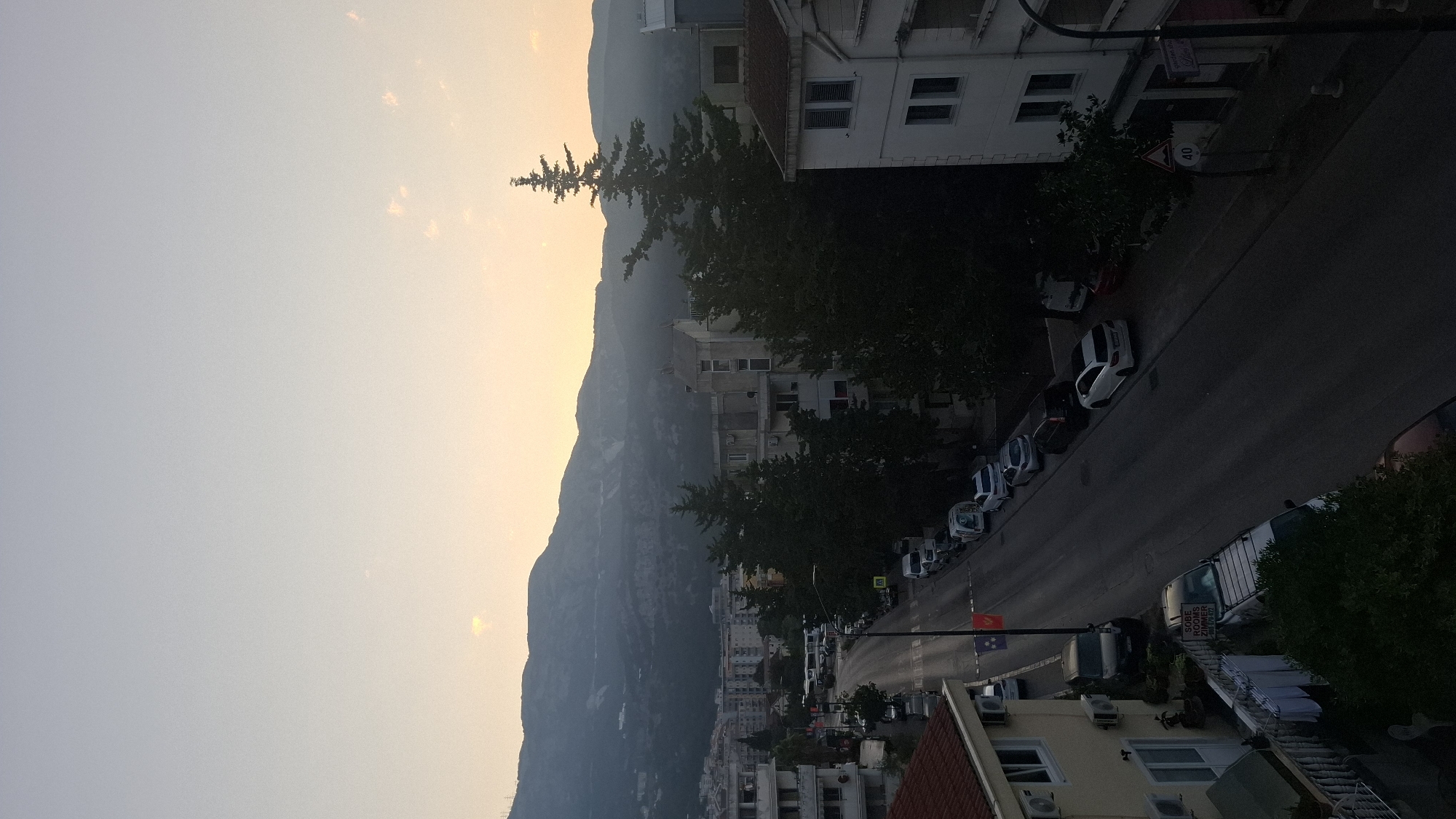 The image captures a street scene in Budva, Montenegro, during twilight or early morning. The sky is a soft gradient of pale orange to light blue, with distant mountains silhouetted against it. The street is lined with multi-story buildings on both sides, with trees and some vegetation present. Several cars are parked along the street and in driveways. There are no people visible in the scene. The buildings appear to be residential or commercial. A speed limit sign showing "40" is visible on the right side of the street. Laundry is hanging on a clothesline in the foreground on the left. The overall impression is a quiet urban environment bathed in soft, natural light.